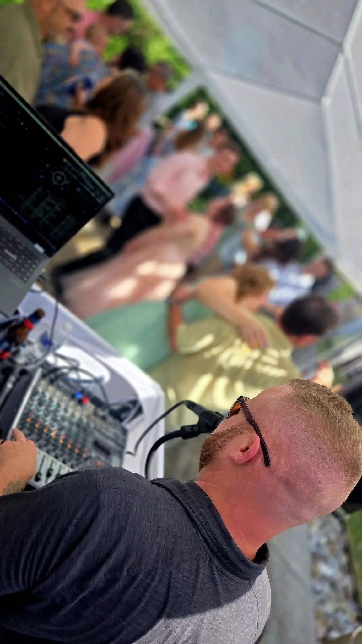 A DJ wearing sunglasses working on sound equipment at an outdoor event with a crowd of people in the background.