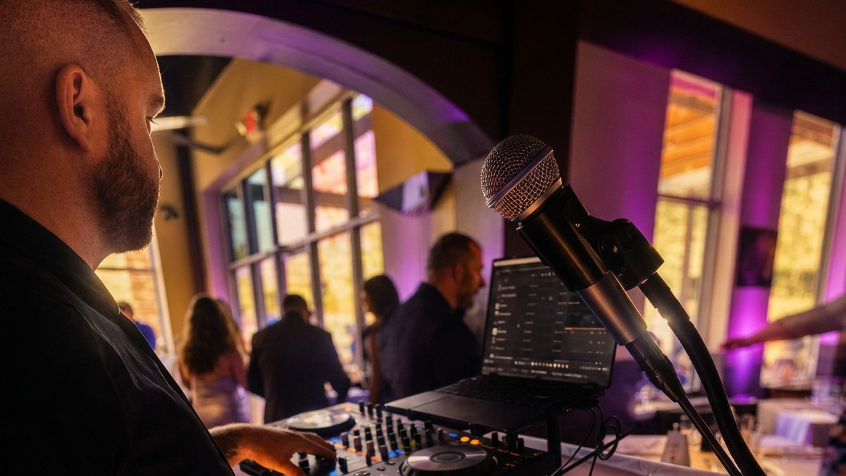 A DJ wearing a black shirt operating a mixer and a laptop at an indoor event with large windows and purple lighting, with people socializing in the background.