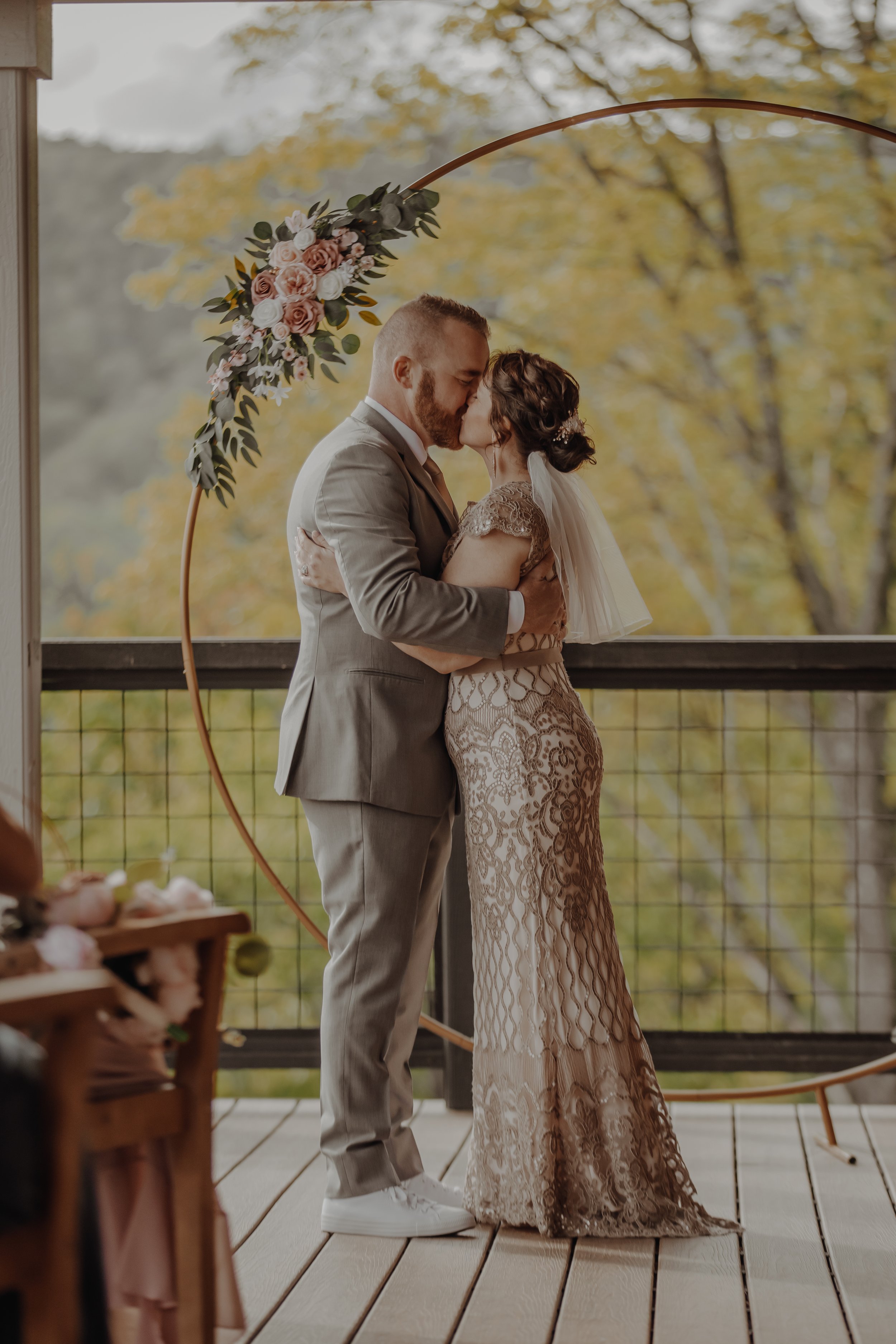 A couple sharing a kiss during their wedding ceremony, standing on a wooden deck with autumn trees in the background, decorated with a floral arch.