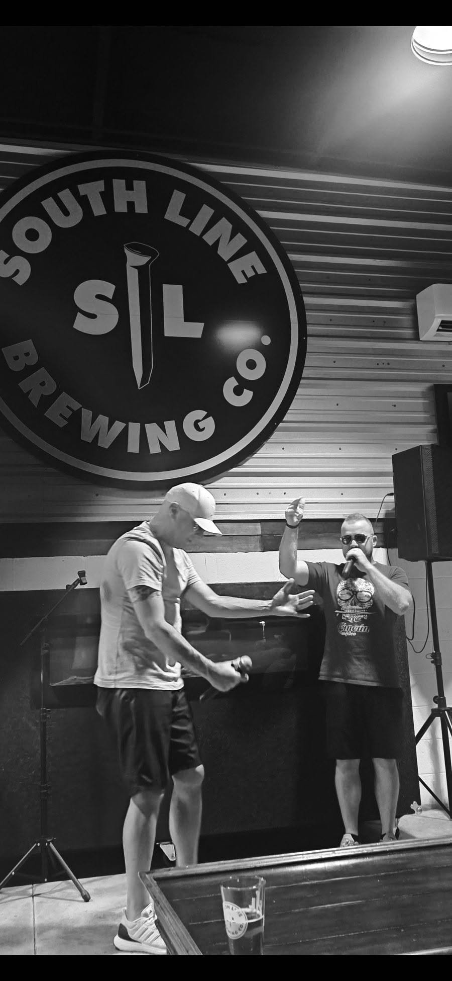 Two men on a stage at South Line Brewing Co. with a large circular sign behind them. One man is wearing a cap and t-shirt, holding a microphone, and reaching out to the other man, who is wearing sunglasses, a graphic t-shirt, and also holding a microphone. The scene appears to be a performance or karaoke night in a brewery.