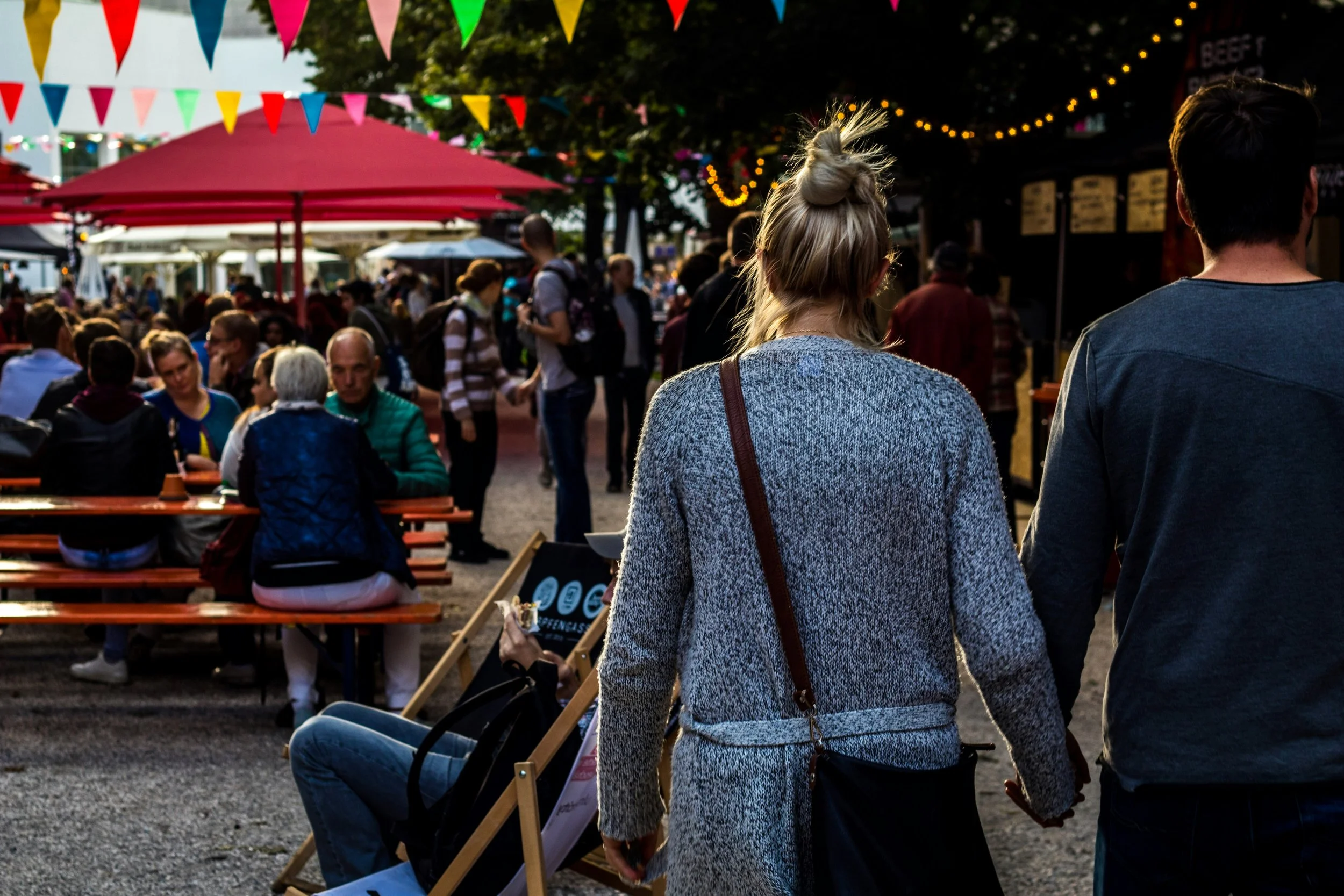 A woman with blonde hair in a bun, wearing a gray sweater, is standing next to a man at an outdoor event. The event has colorful triangular pennant banners, a pink canopy, and string lights. People are gathered around tables, and some are standing or walking in the background.