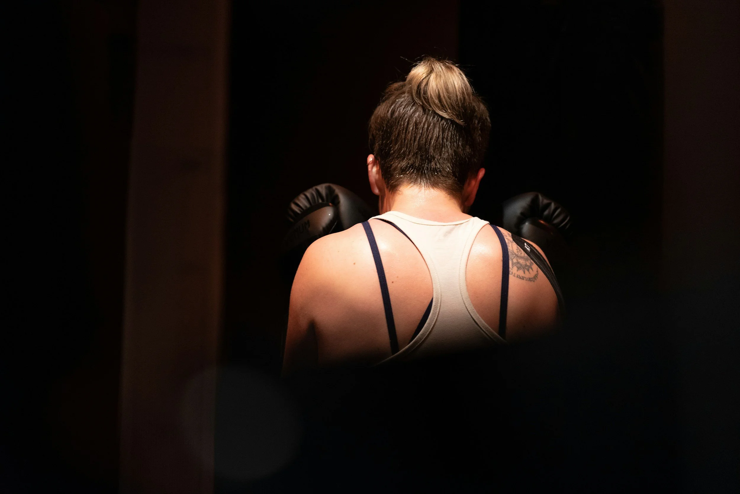 A woman with short brown hair is standing with her back to the camera in a dimly lit environment, wearing a white tank top with black straps and boxing gloves.