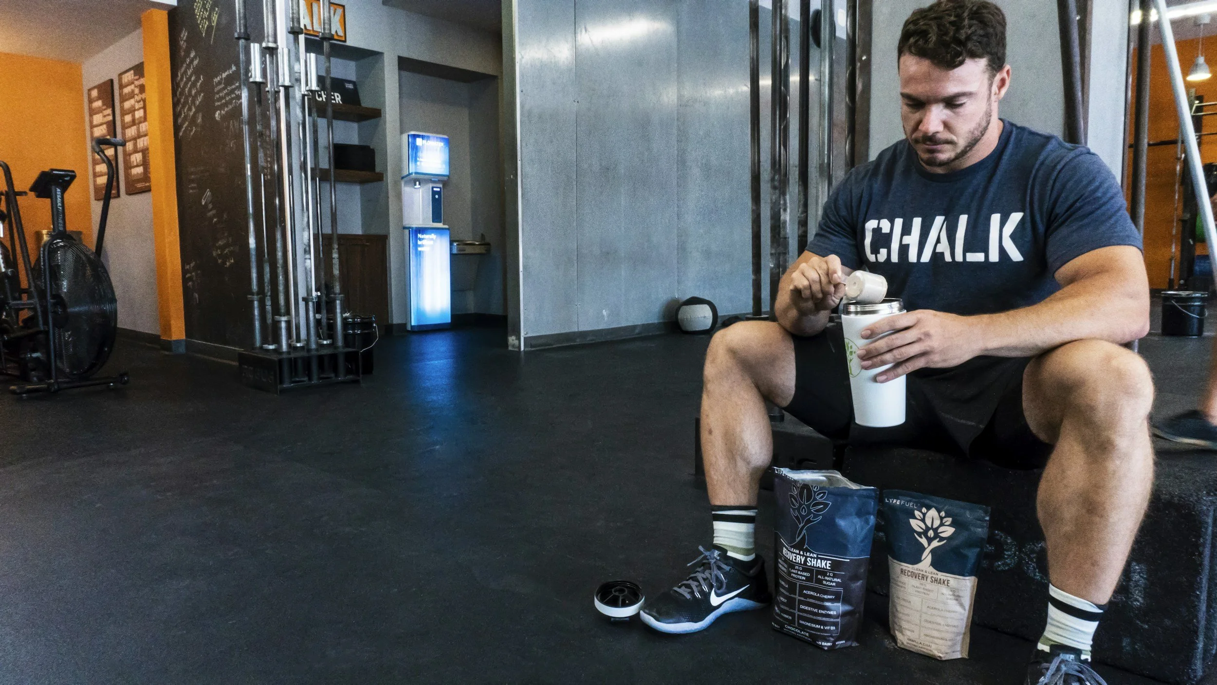 A man sitting in a gym, wearing a blue shirt with 'CHALK' written on it. He is opening a container while seated on a workout bench, with workout equipment and a chalk bag nearby, and gym walls and flooring in the background.