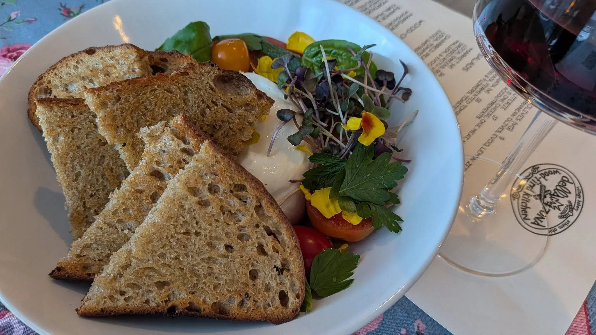 Sourdough bread slices, mixed greens salad with cherry tomatoes, and a glass of red wine on a table.