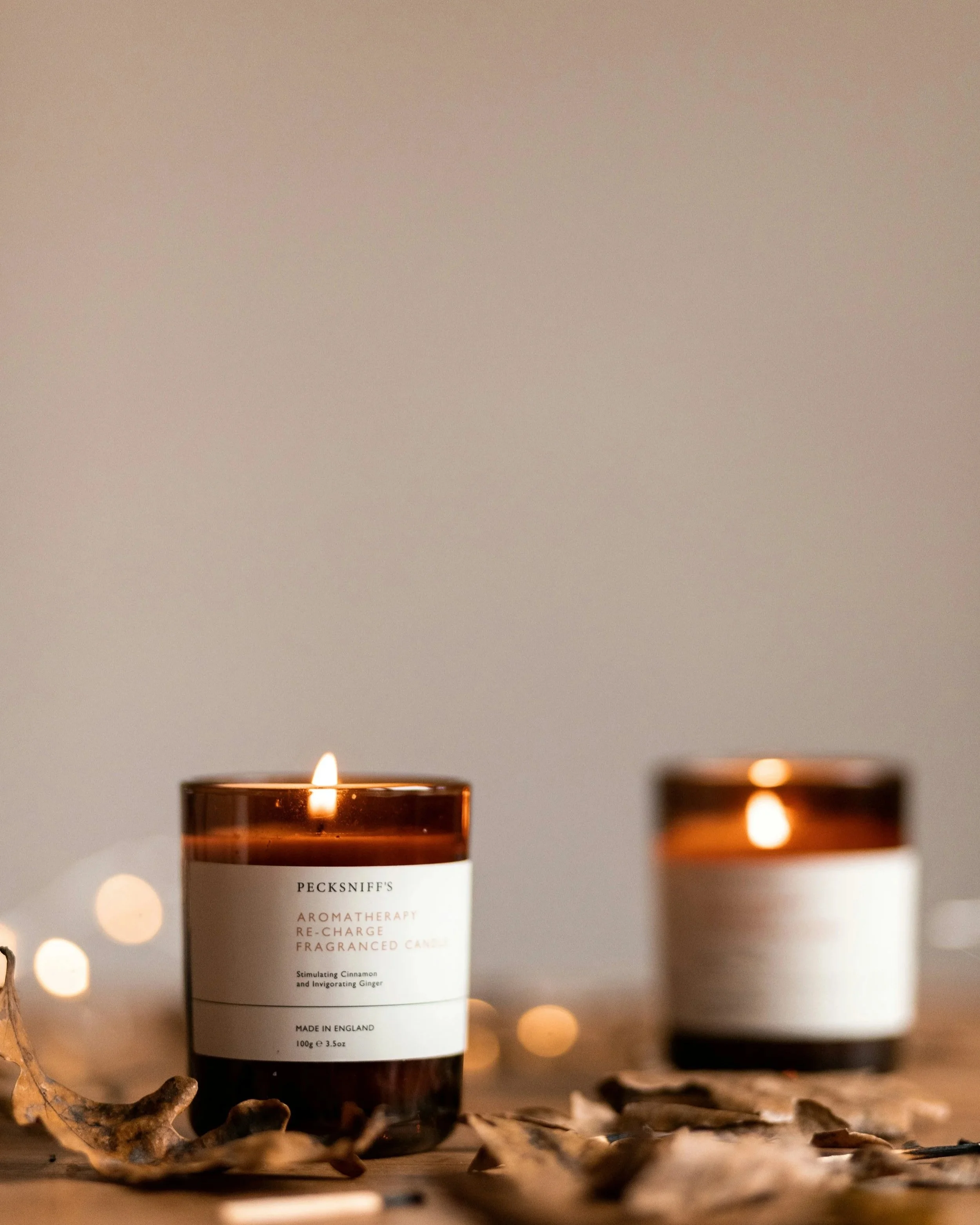 A lit aromatherapy candle labeled 'Pecksniff's' on a wooden surface with dried leaves around it and additional candles blurred in the background.