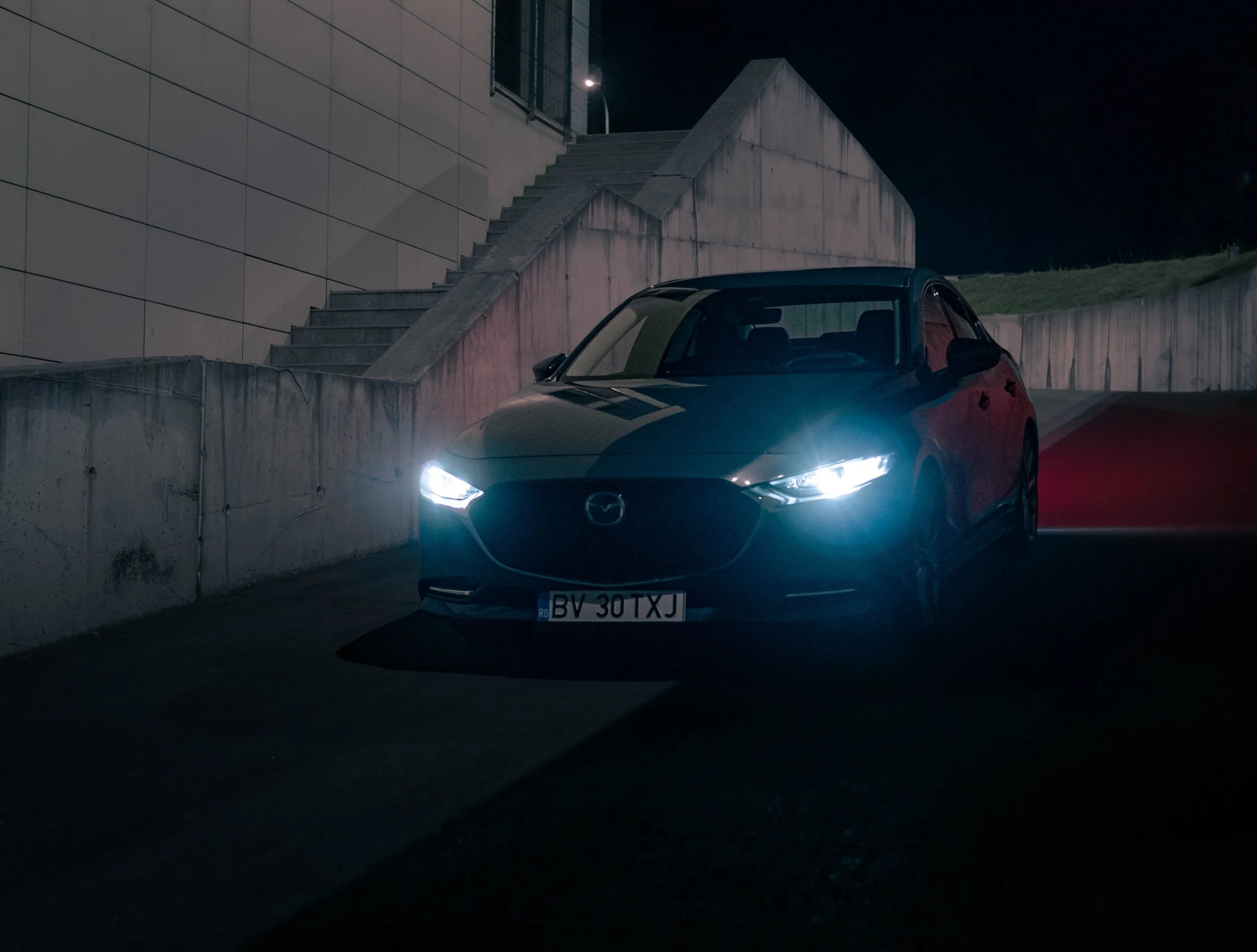 A dark street scene at night with a modern black Mazda car with headlights on, parked near a concrete wall and stairs, in front of a building with a staircase leading up to an opening, and a grassy hill in the background.