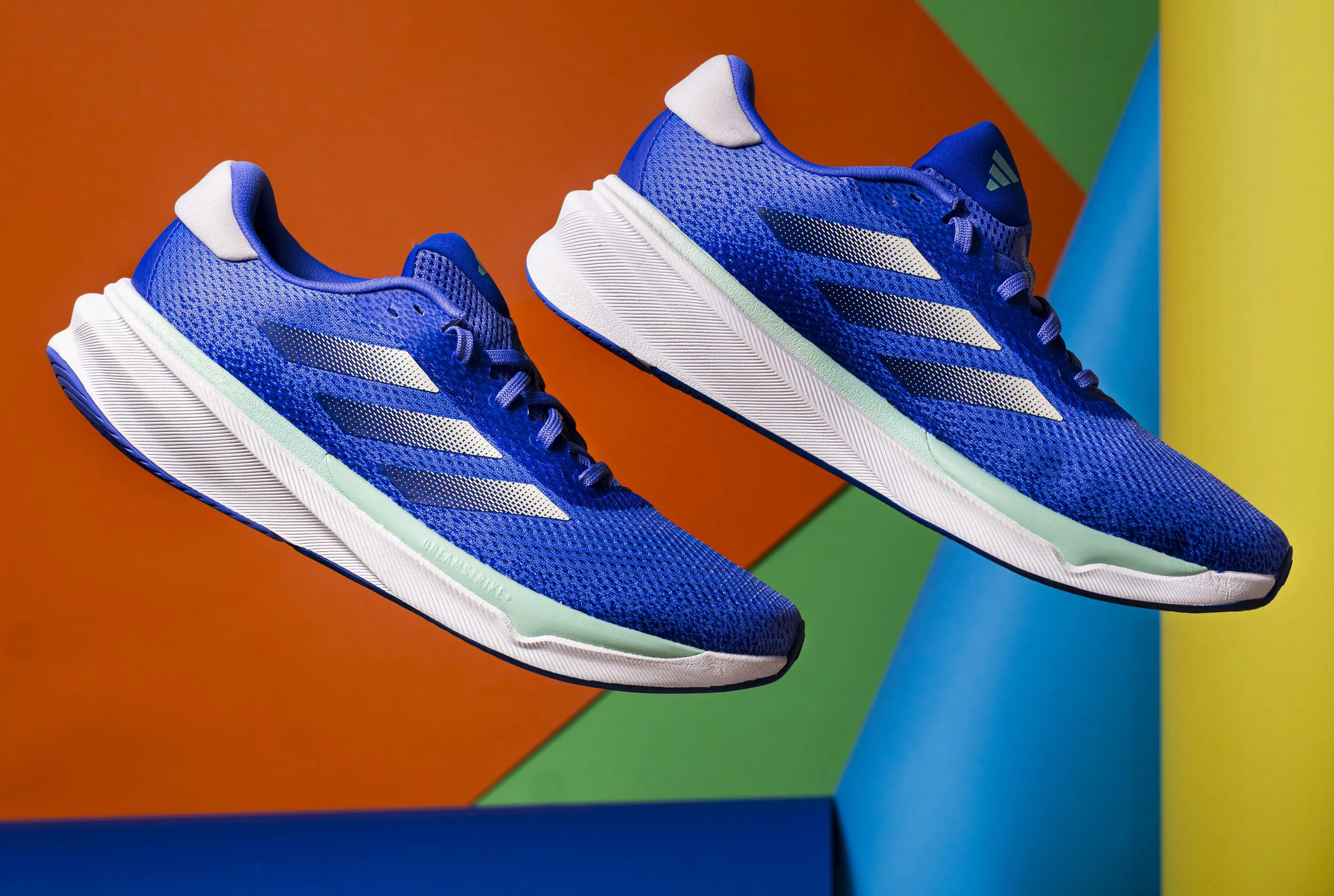 A pair of blue athletic running shoes with white soles and striped accents, placed against a colorful geometric background with orange, green, and blue sections.