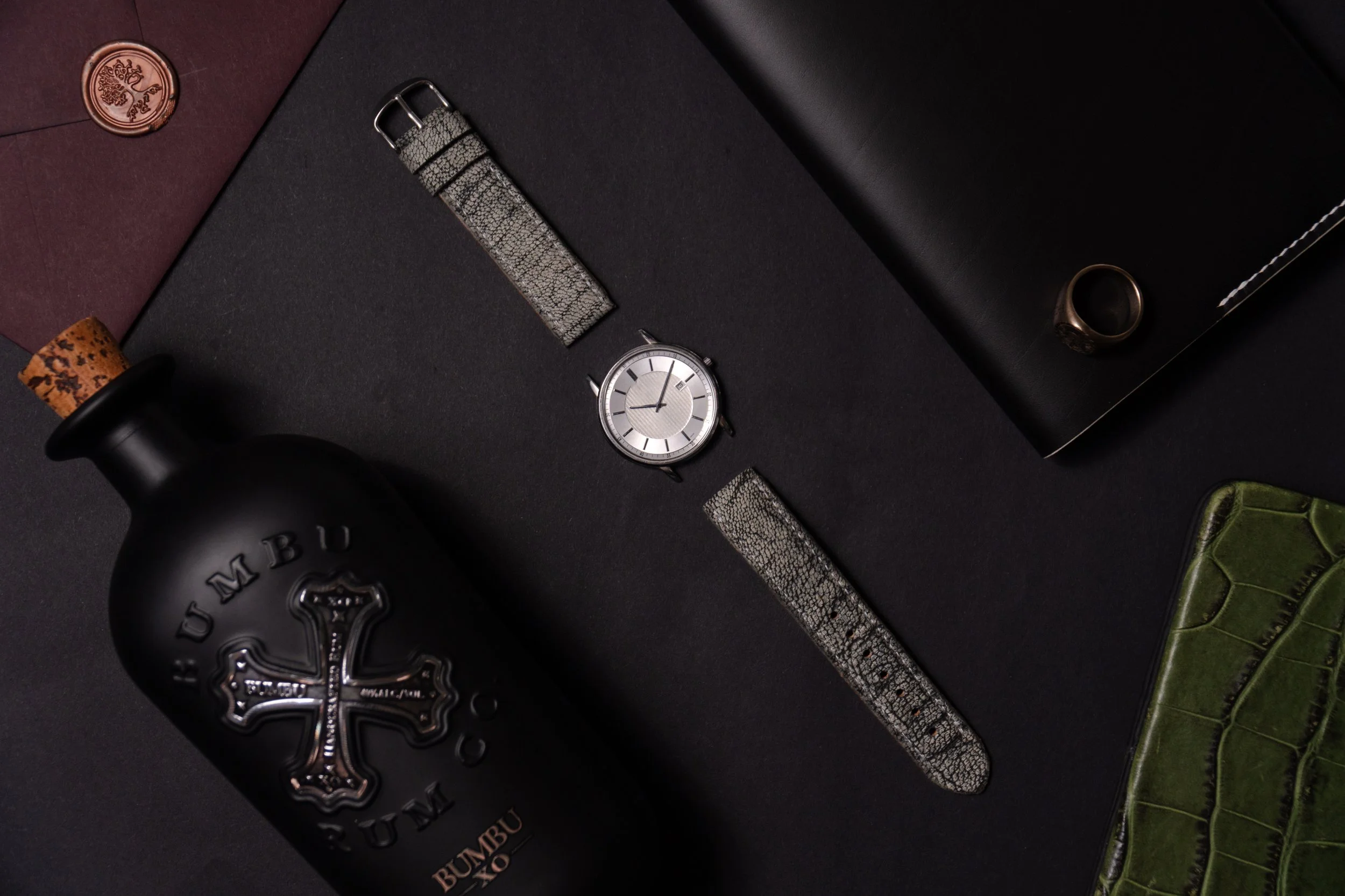 Flat lay of a black bottle with a cork stopper, a silver wristwatch, a patterned silver wristband, a black rectangular box, a silver ring, and a green wallet on a black background.