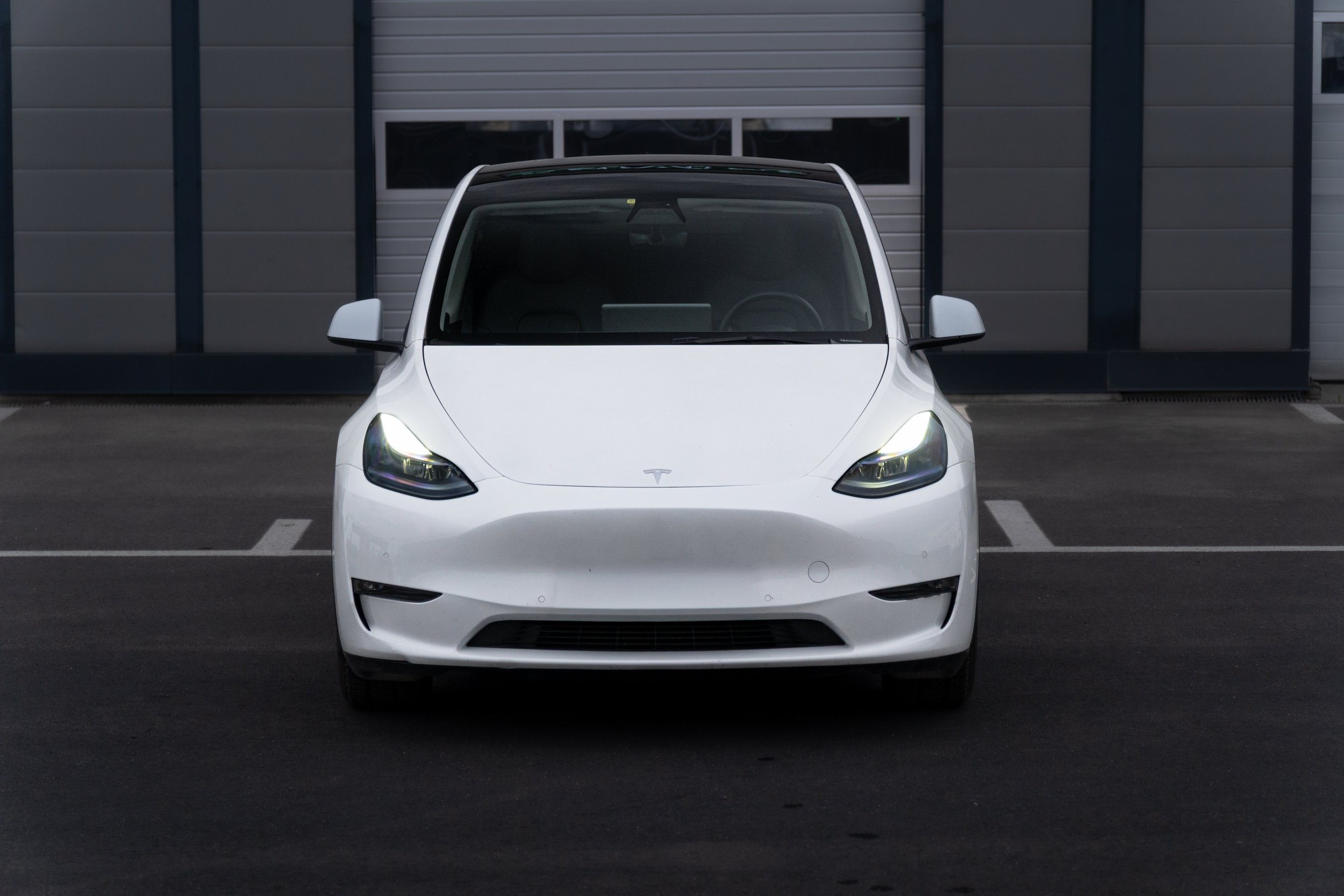 Front view of a white Tesla car parked in a parking lot in front of a gray building with windows.