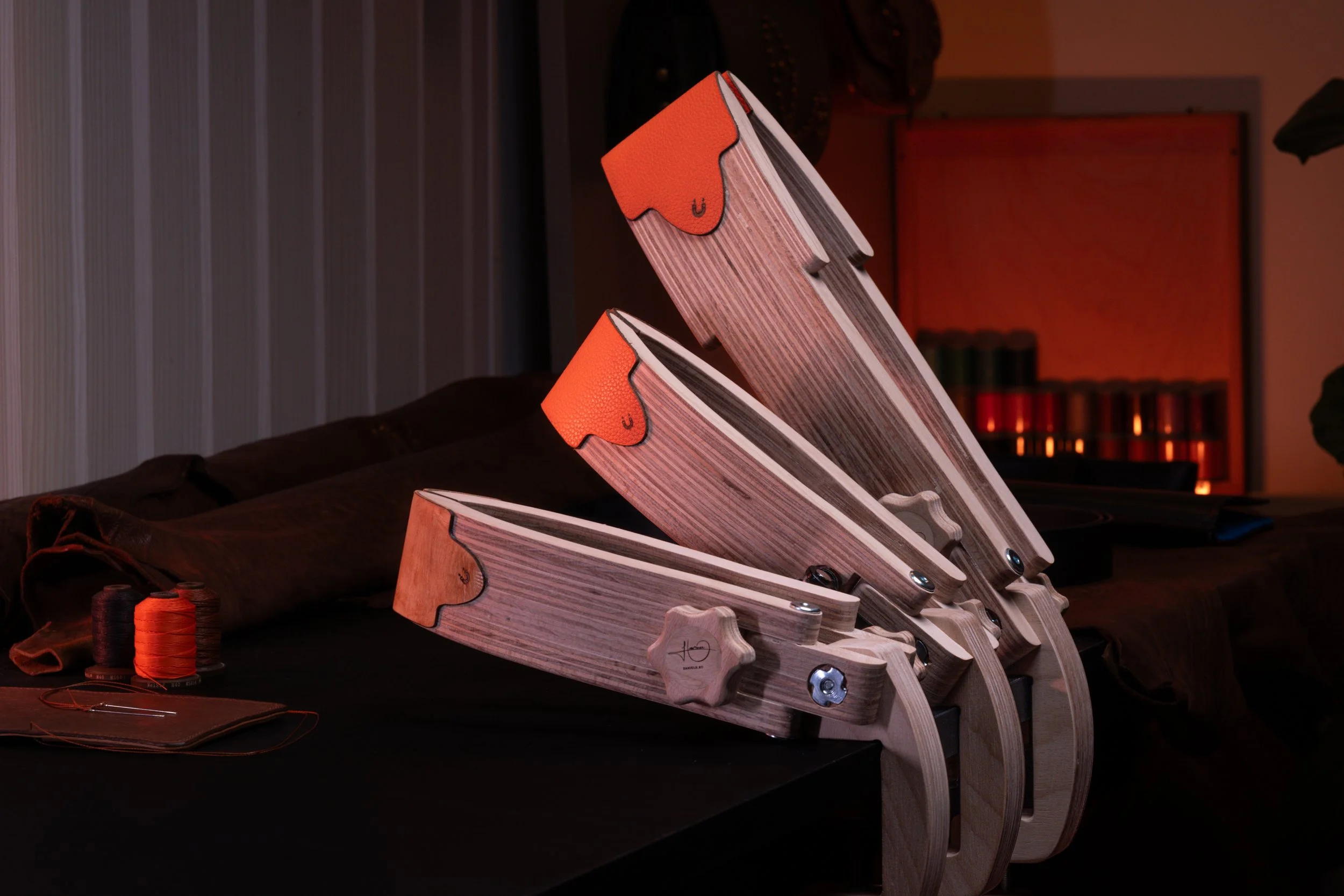 A set of wooden skateboards with orange grips are arranged on a black surface in a dimly lit room. In the background, there are spools of black and orange thread, a leather item, and a bookshelf with orange lighting.