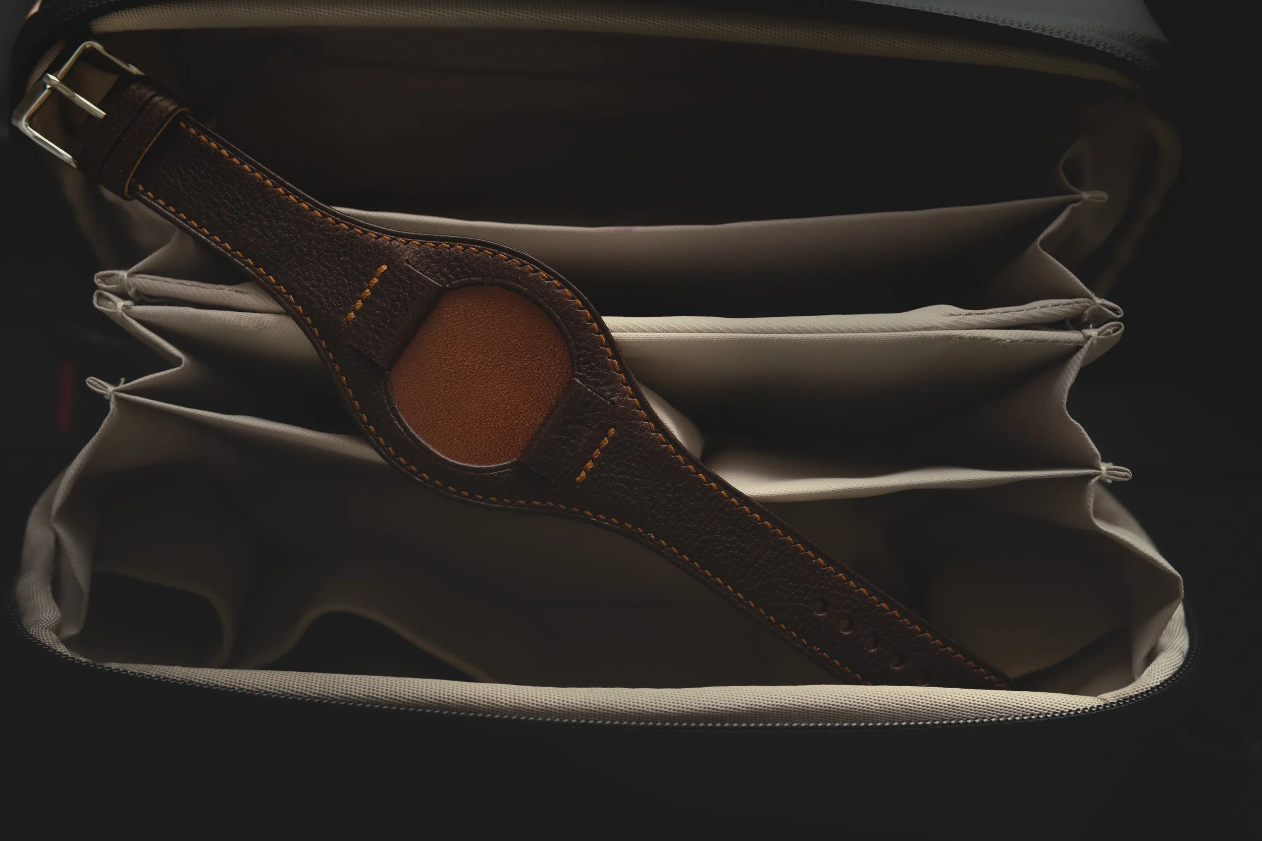 Inside of a bag with a beige fabric lining, a brown leather belt with orange stitching, and multiple zipped compartments.