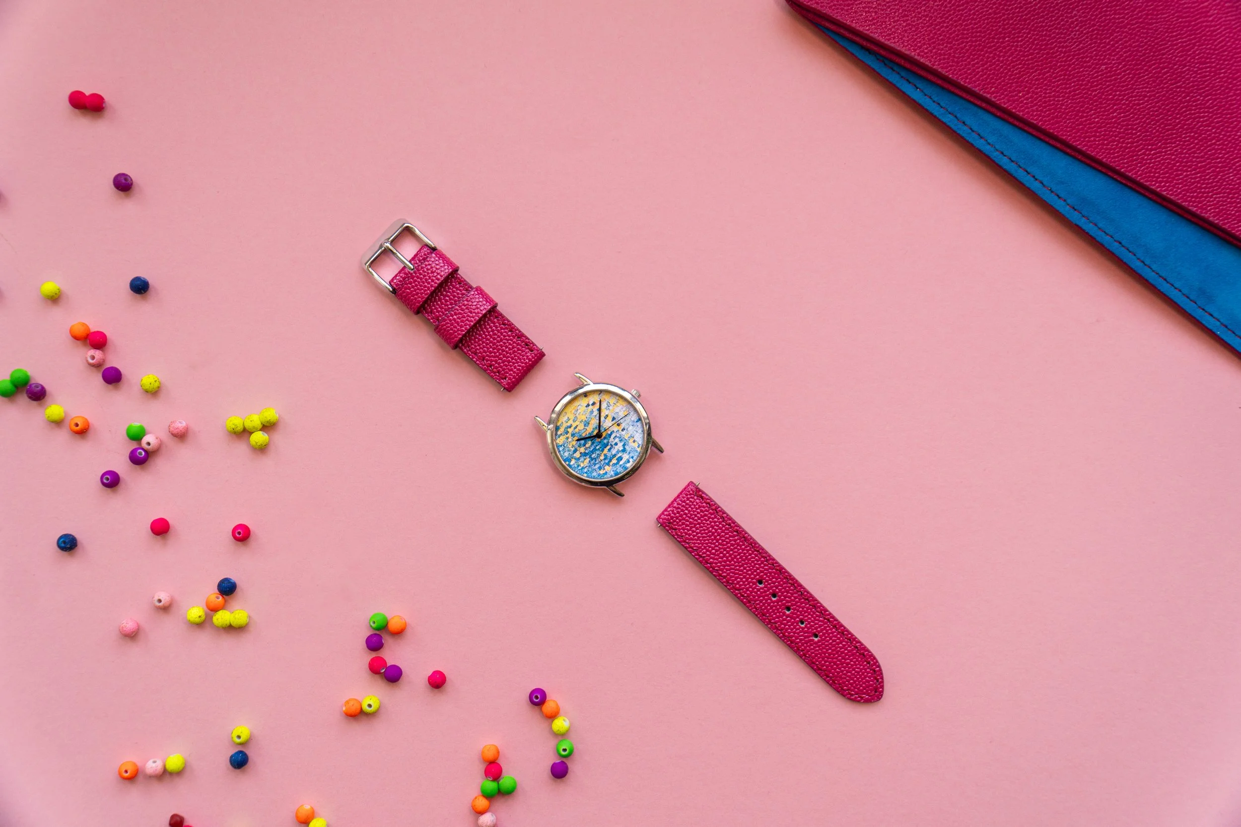 Colorful array of small round beads scattered on pink background, with a pink watch with a colorful face, pink strap, and two pink books with textured covers in the top right corner.