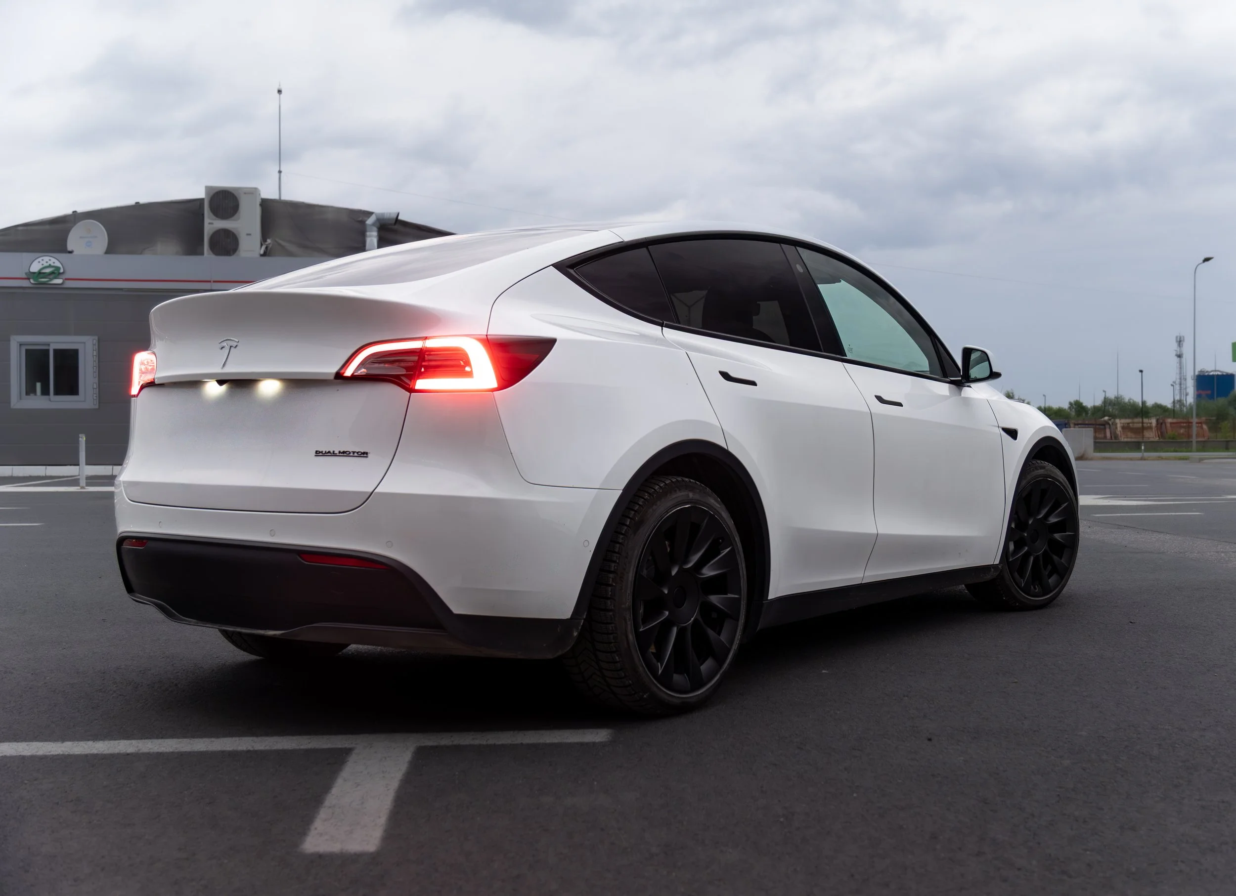 A white Tesla Model Y electric vehicle parked in an outdoor parking lot under cloudy sky, with visible tail lights, black rims, and a 'Dual Motor' badge.