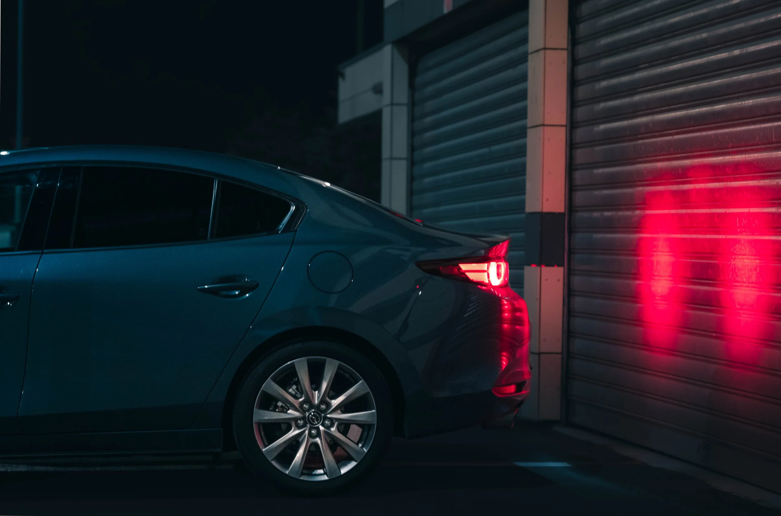 A dark-colored sedan parked near a metal roll-up door with red taillights illuminated at night.