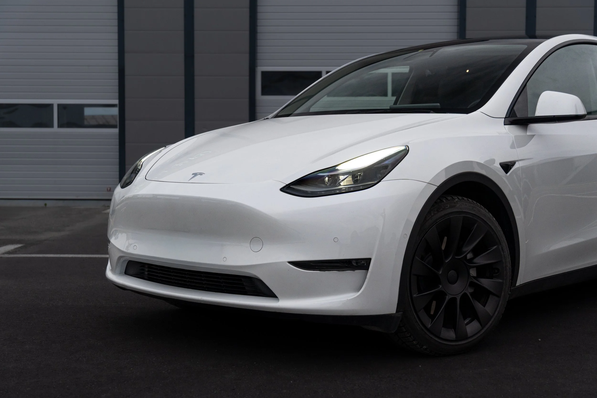 A white Tesla Model 3 electric car parked outside a modern building with gray panels and large windows.