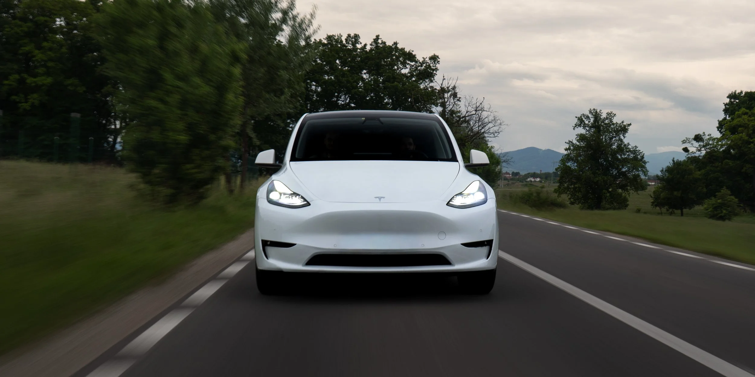 A white Tesla electric vehicle driving on a scenic two-lane road surrounded by greenery and trees, with mountains in the distance under a cloudy sky.