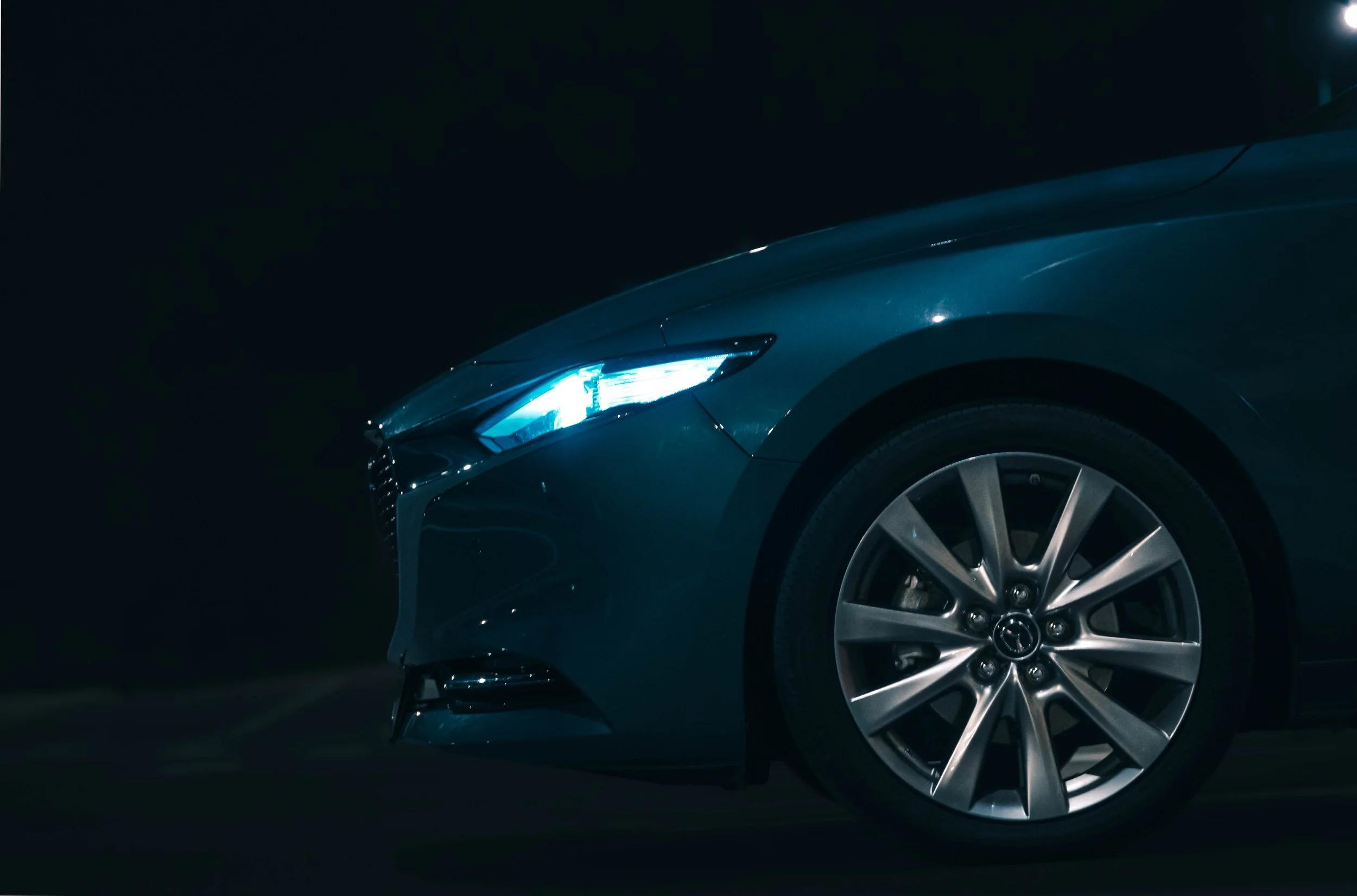 Front part of a dark blue or black modern car with illuminated headlights parked on a reflective surface in a dark environment.