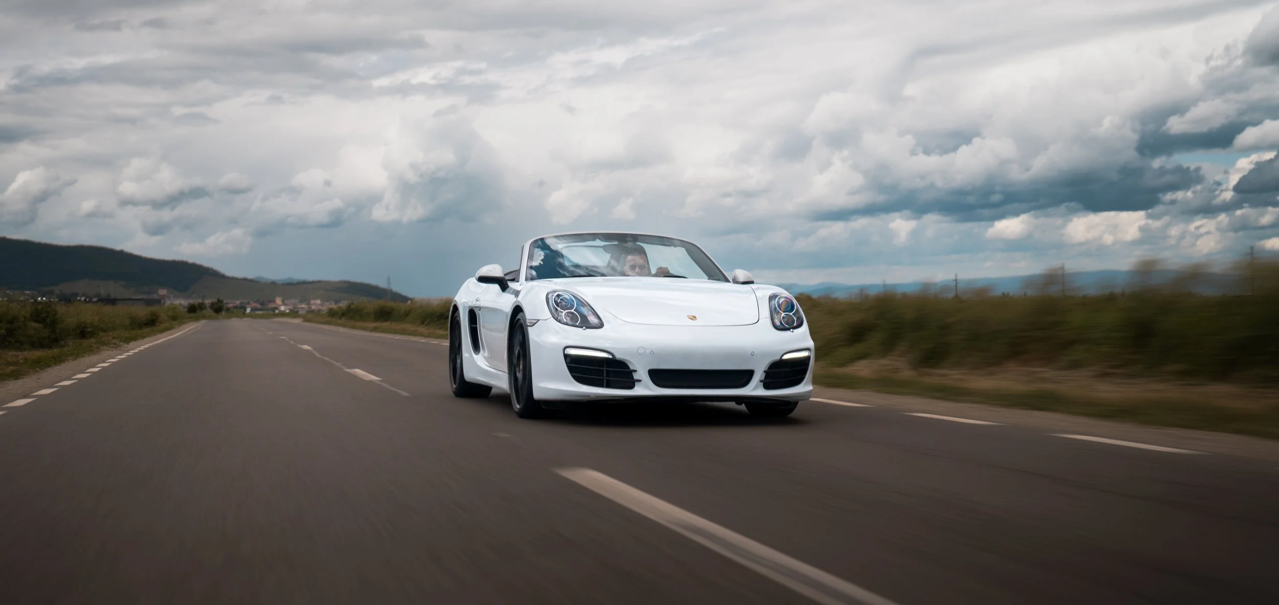 A white Porsche sports car driving on an open road with green fields and hills in the background under a cloudy sky.