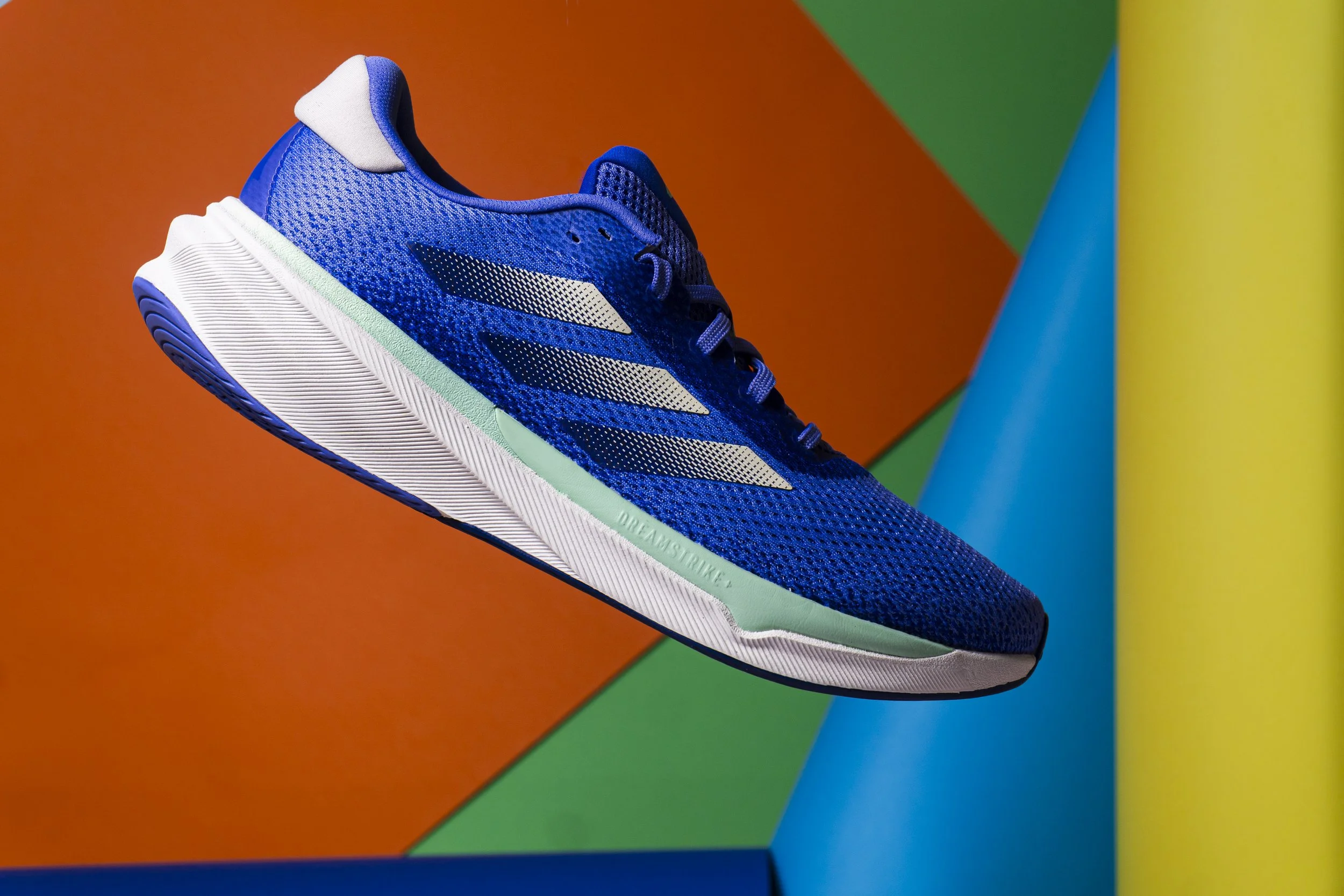 Close-up of a blue running shoe with white and light green accents, set against a background of colorful geometric shapes in orange, green, yellow, and blue.