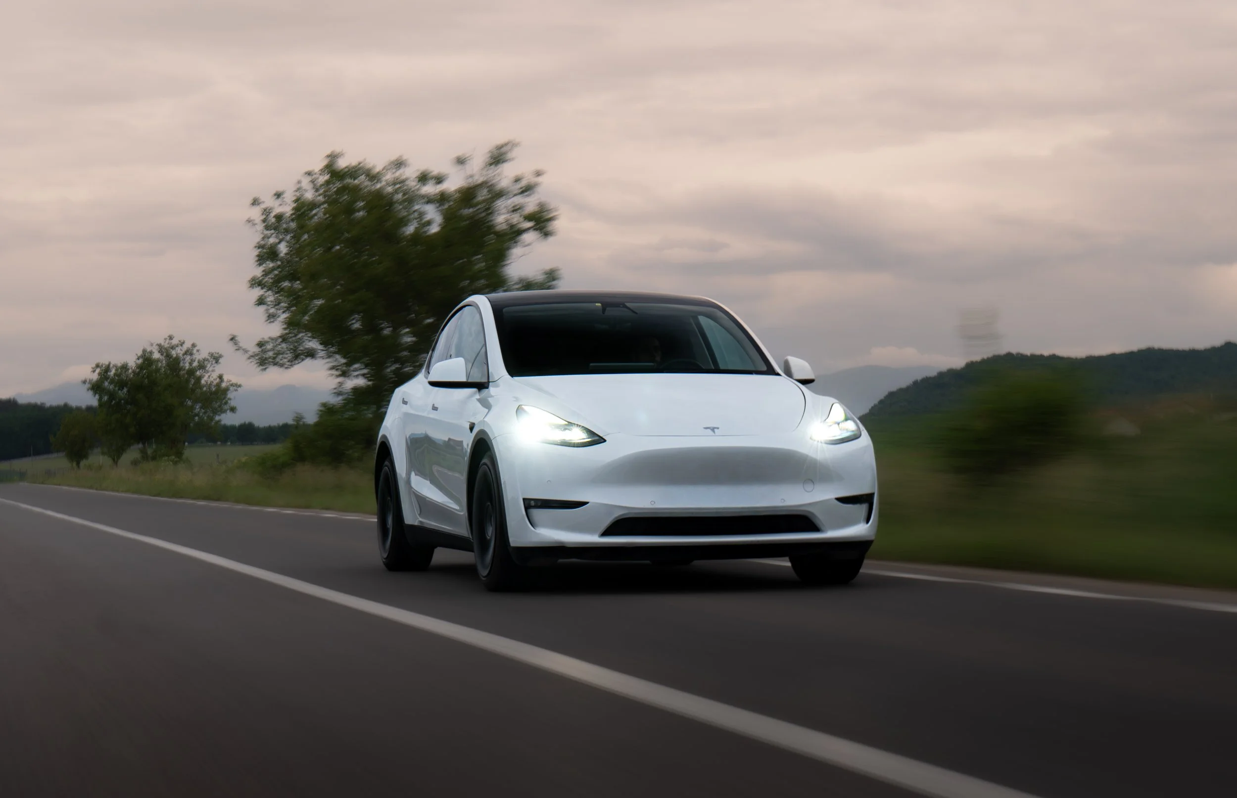 A white Tesla Model Y electric vehicle driving on a two-lane rural road with green trees and rolling hills in the background under cloudy sky.