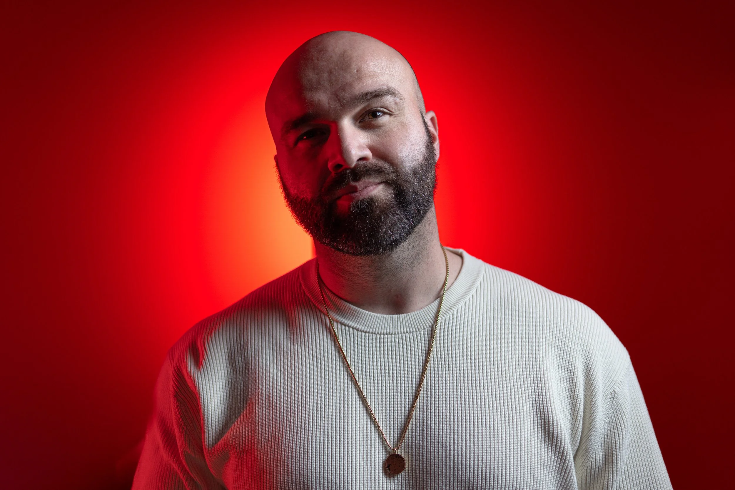 A portrait of a bald man with a beard wearing a white ribbed sweater and a necklace with a pendant, standing against a red background.