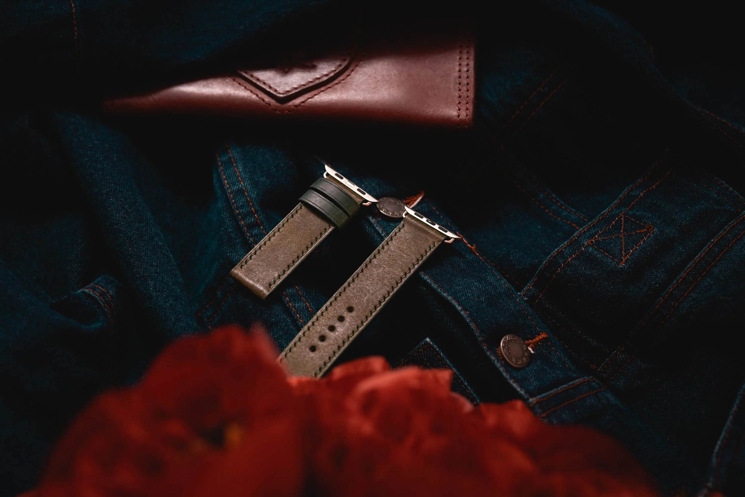 Close-up of a brown leather wallet and gray watch strap on dark blue denim jeans with orange stitching, partially covered by a dark fabric, with red blurred foreground.