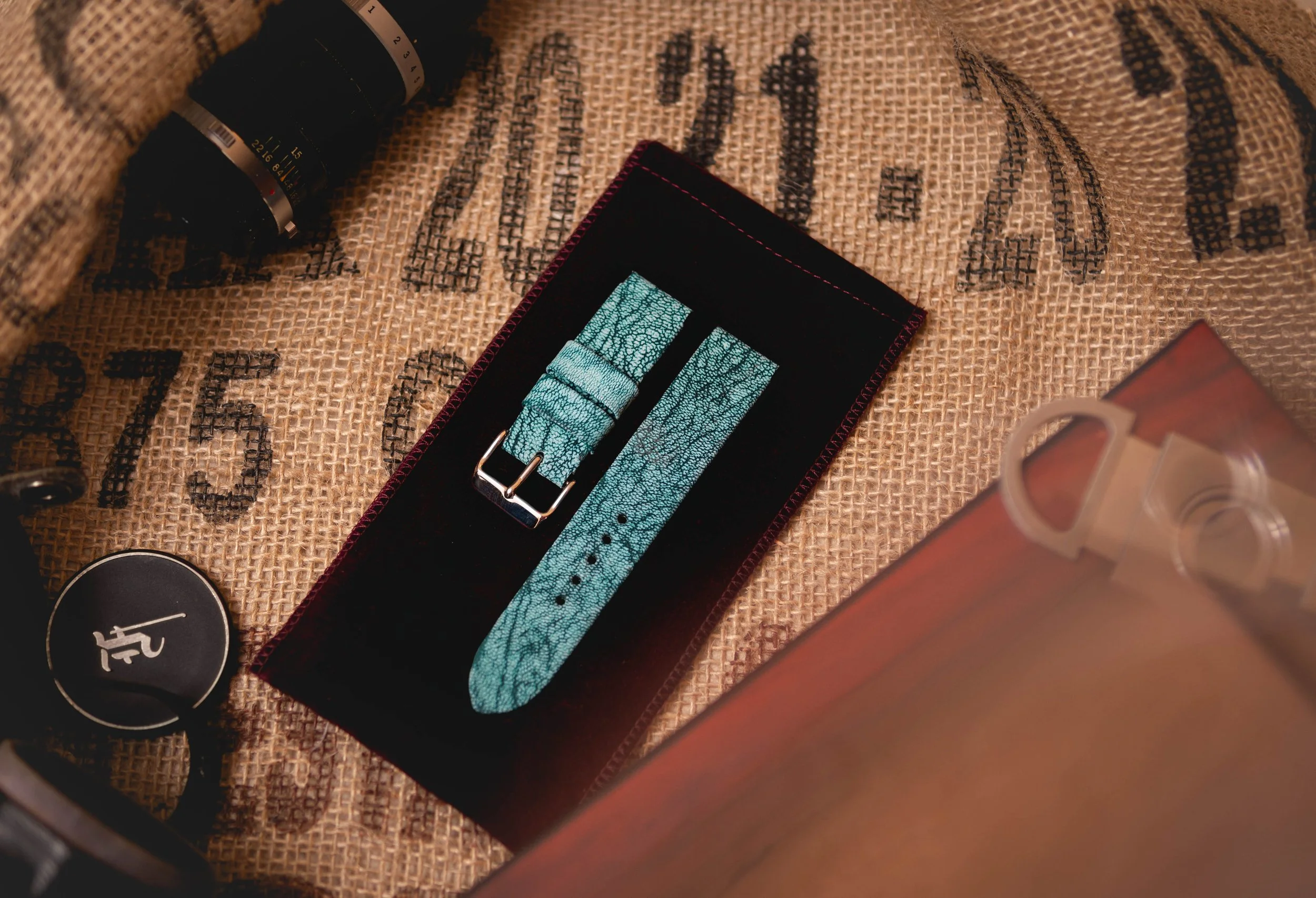 A burlap sack with black printed text, a black-framed watch with a turquoise strap, a black container with a white logo, and a partially visible cardboard box.