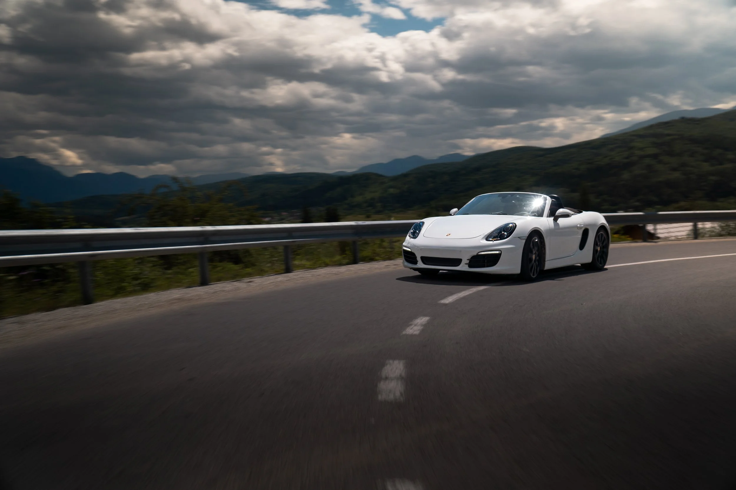 A white convertible sports car driving on a highway through mountainous terrain with cloudy skies overhead.