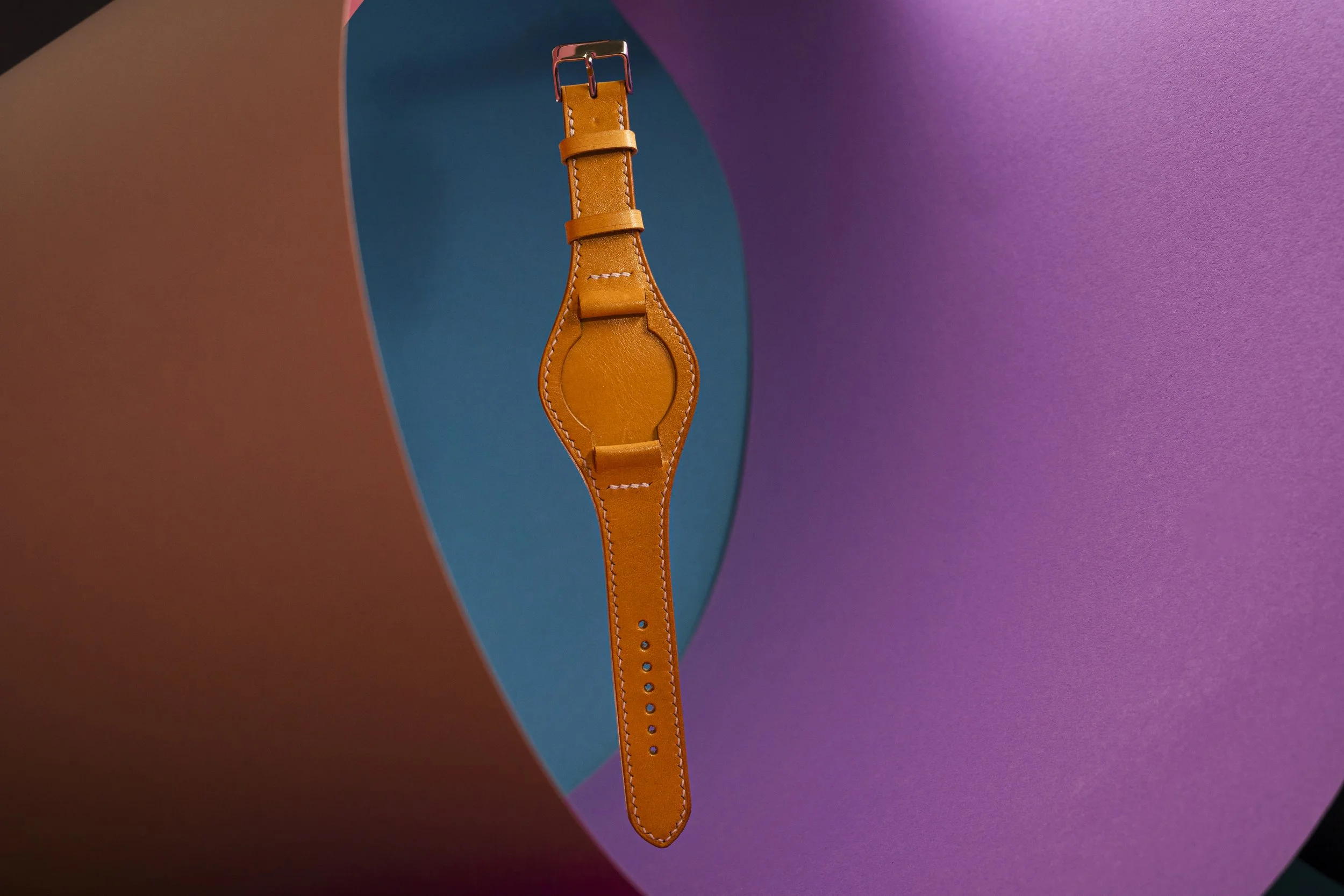 Orange leather wristwatch strap hanging vertically against colorful abstract background with shades of pink, blue, purple, and brown.