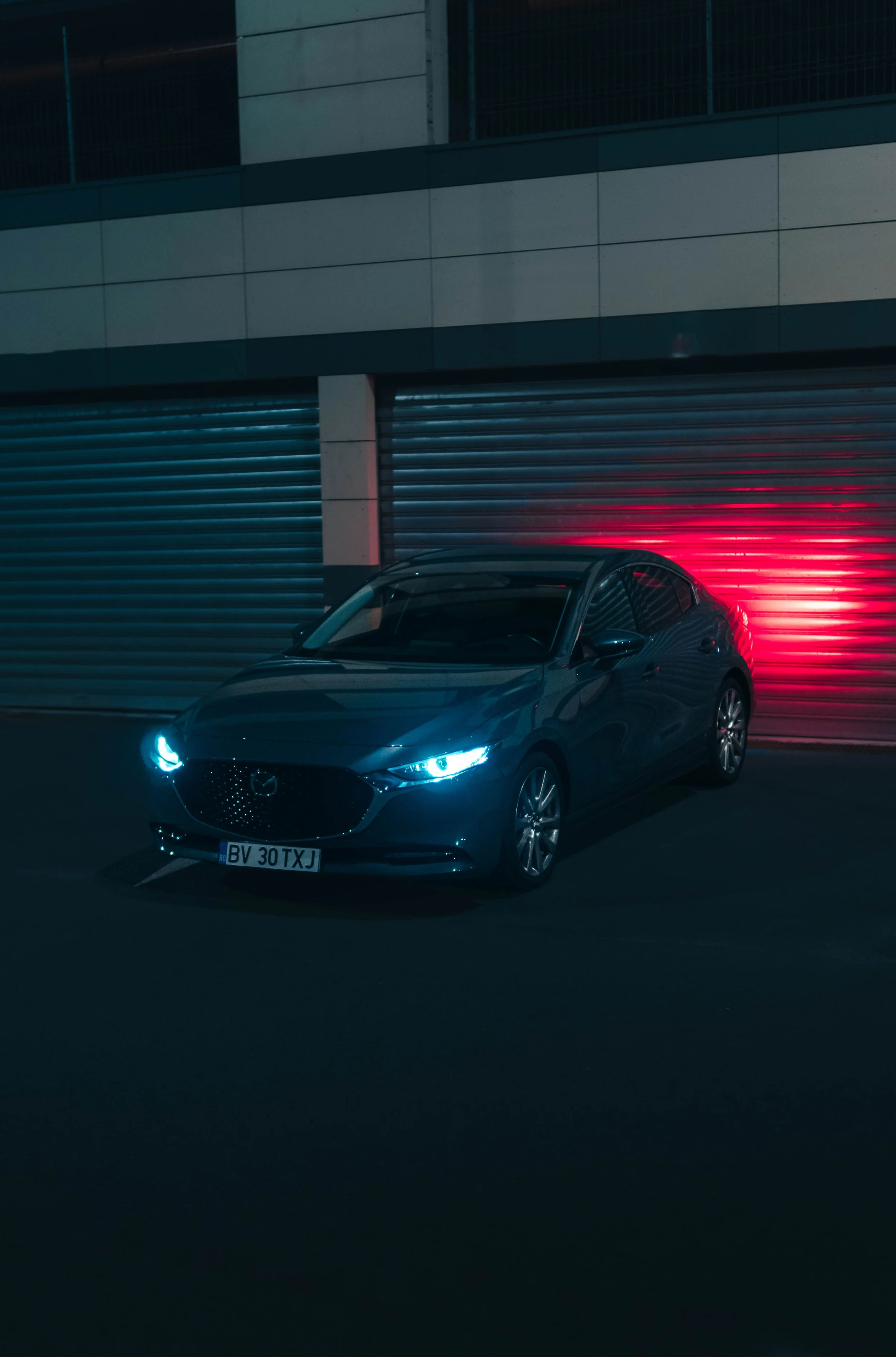 A gray Mazda car with blue and red headlights parked in front of a closed, metal shutter with a red light reflecting on it.