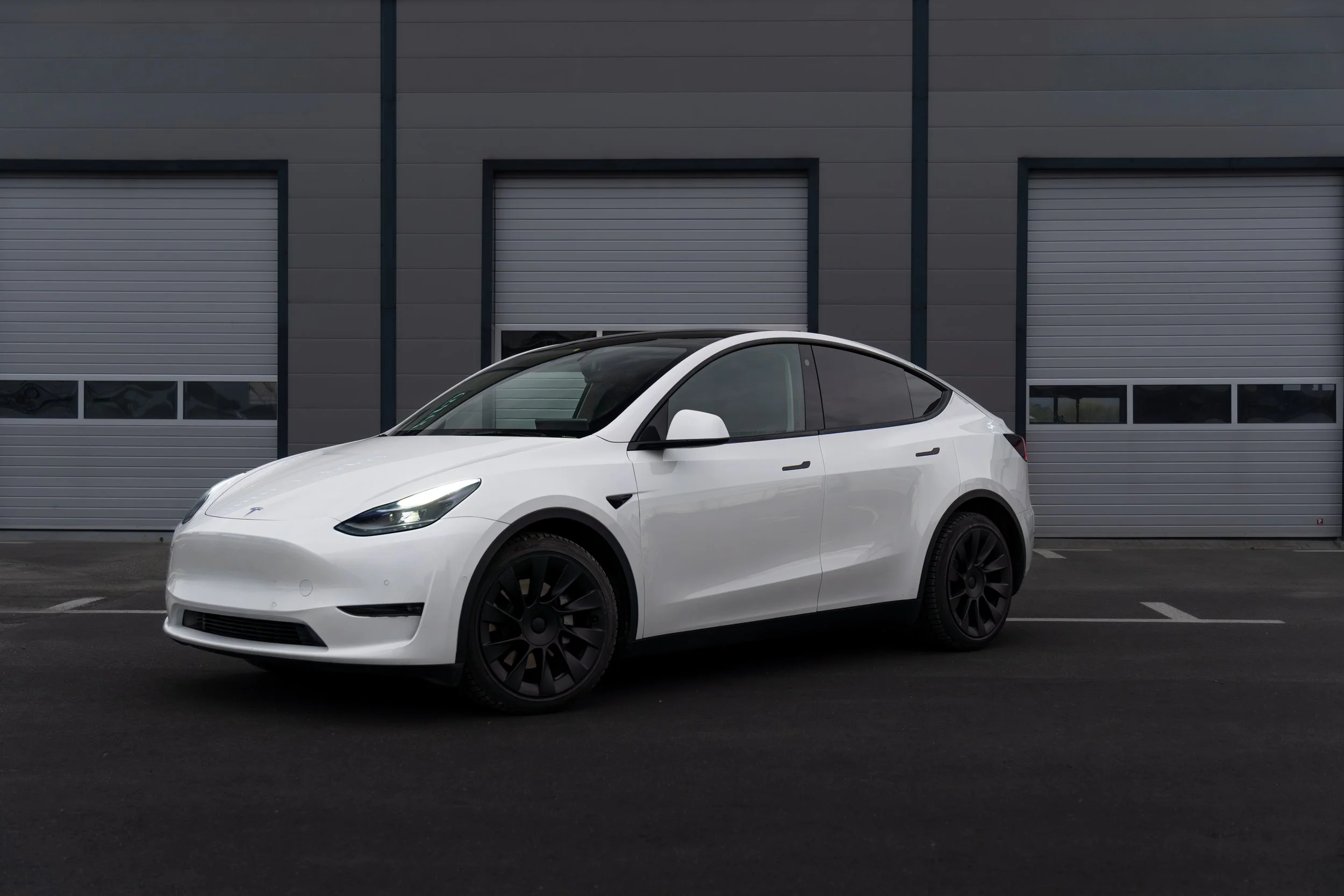 White Tesla parked in front of a gray industrial building with multiple garage doors.
