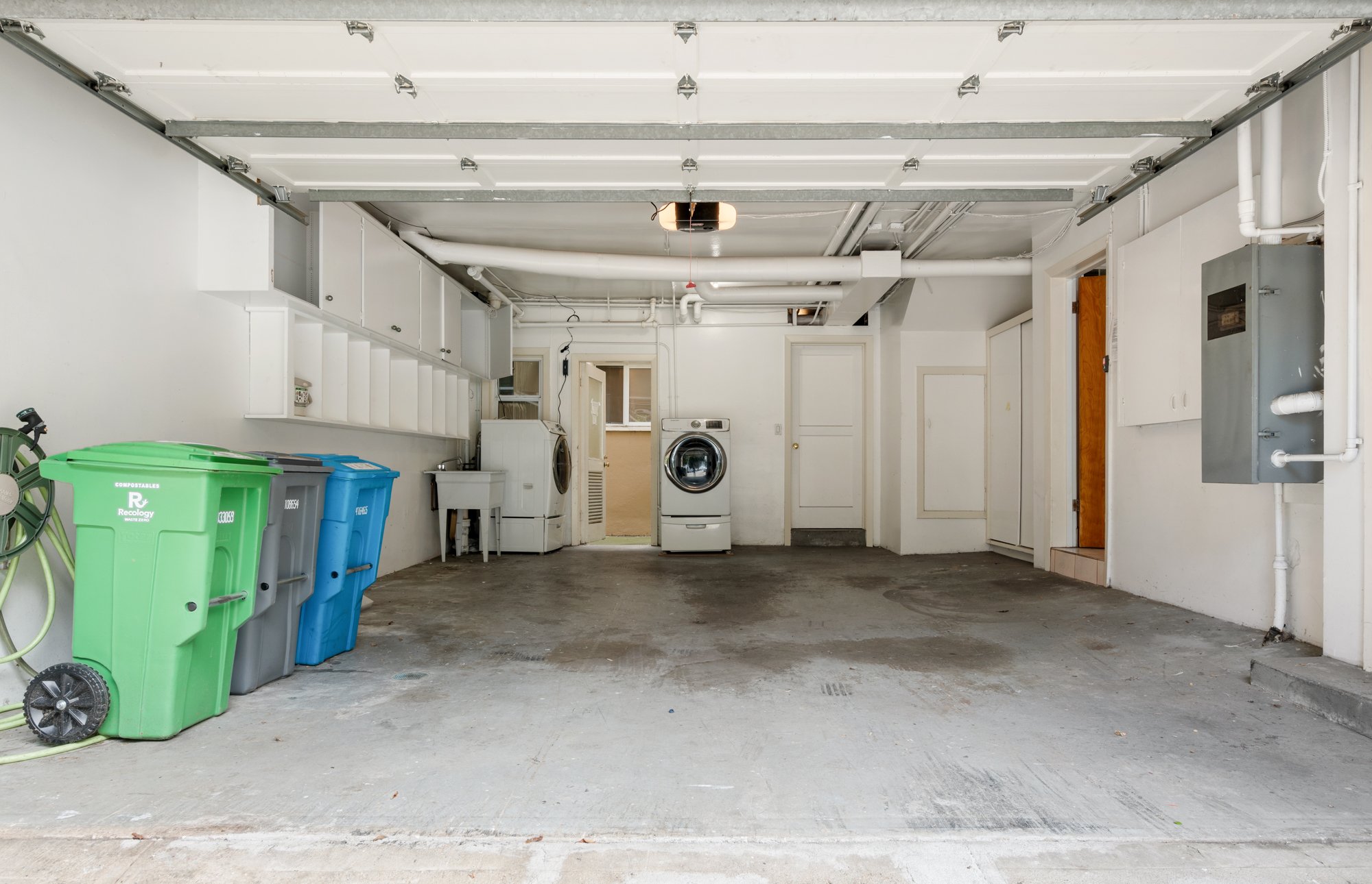A garage with a concrete floor, three trash bins in green, gray, and blue, a garden hose, a laundry sink, a washing machine, a dryer, and a utility sink, with an open door leading outside.
