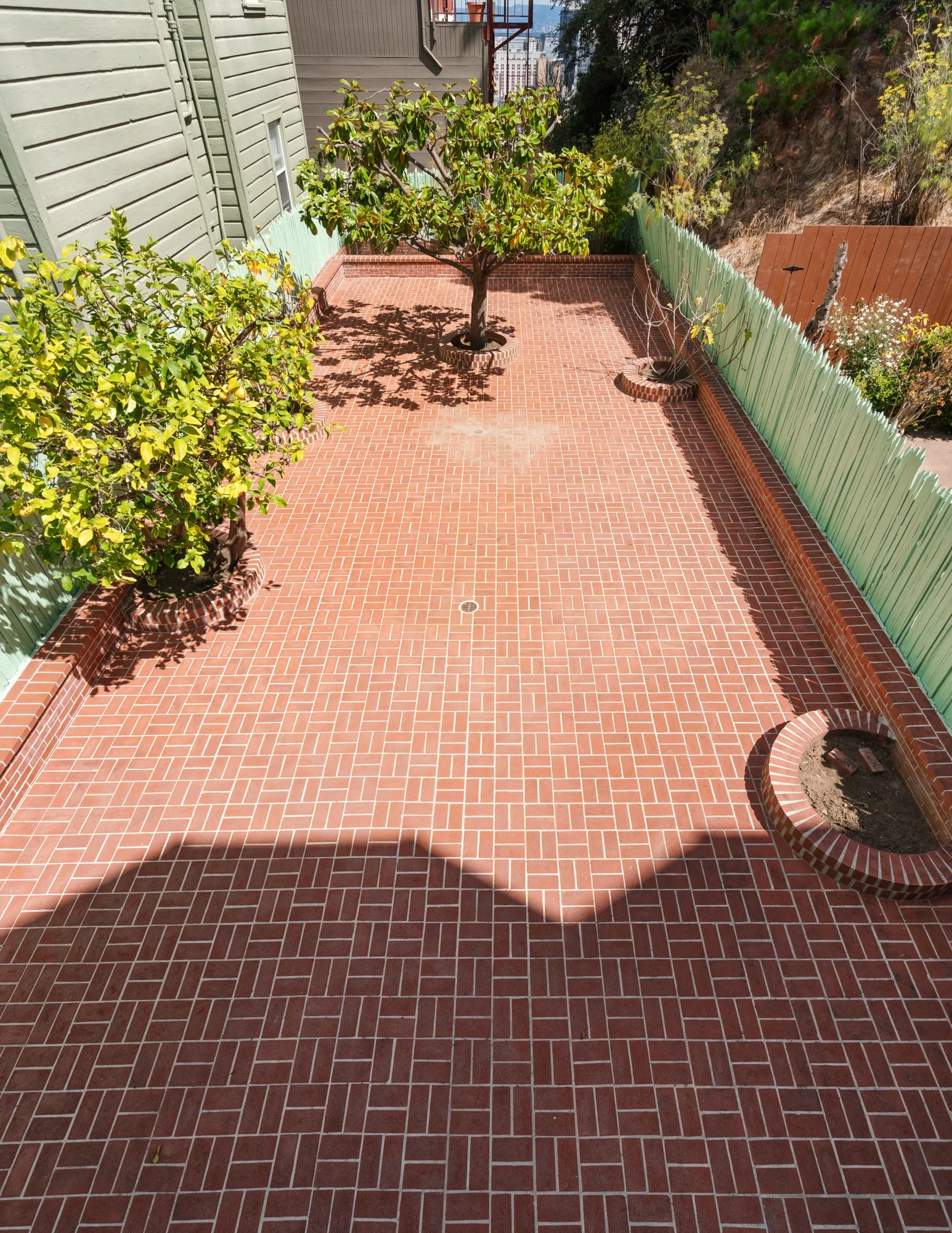 A small backyard patio with red brick flooring, green wooden fence, a tree in the center, and two potted plants on the side. Part of a house and neighboring buildings are visible in the background.
