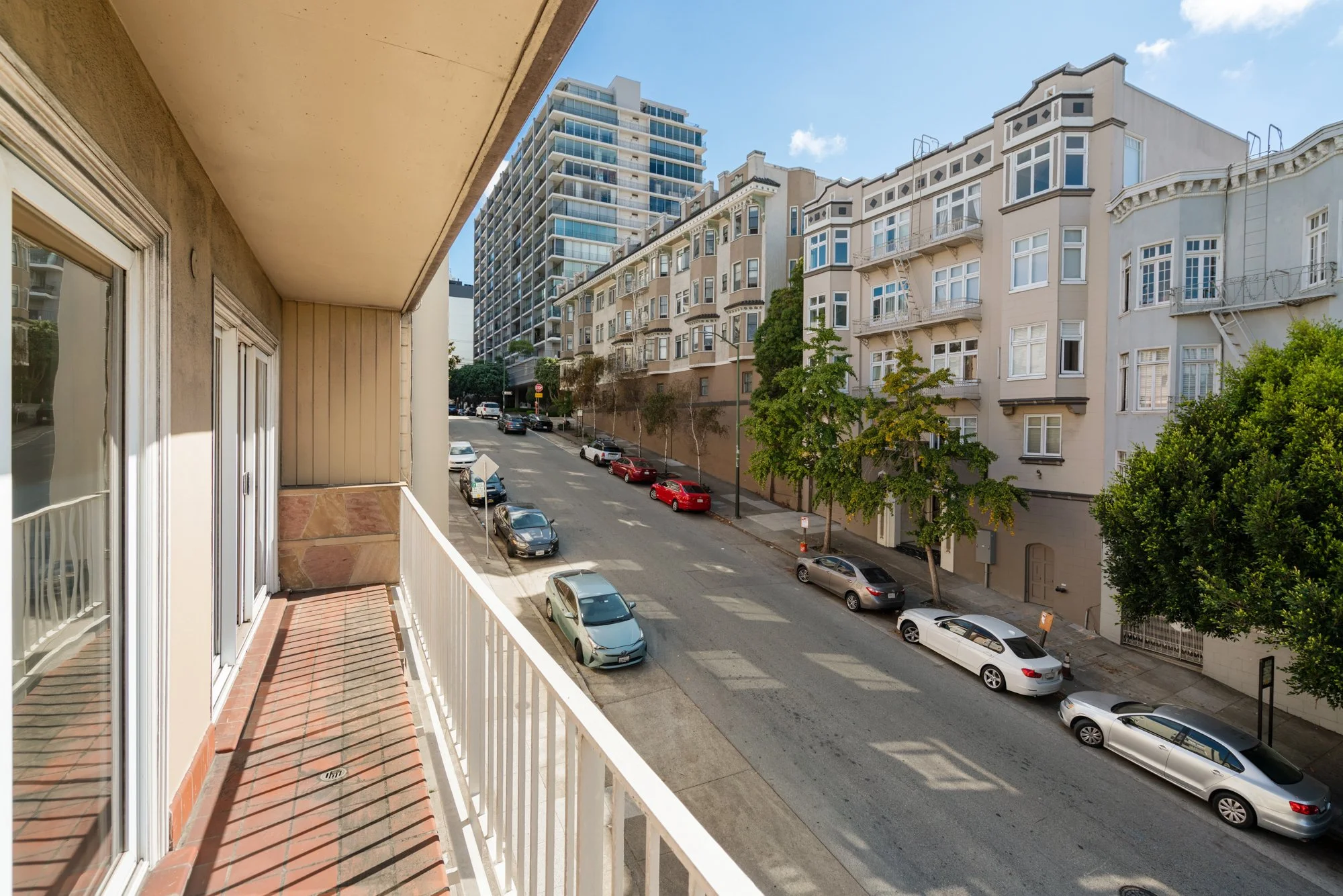 View from a balcony of an urban street with parked cars, trees, and tall buildings under a clear blue sky.