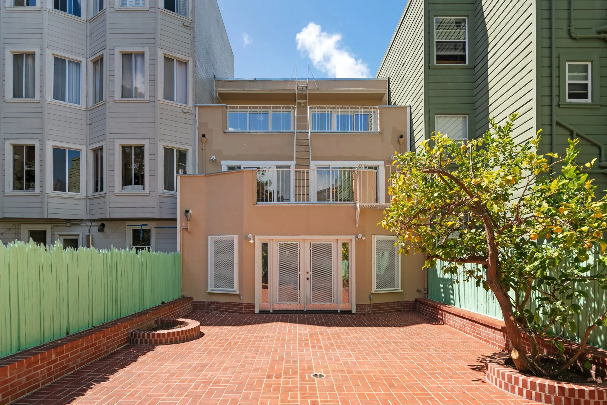 View of a backyard patio with red brick paving, a small lemon tree, green wooden fence on the left, beige and green apartment buildings in the background, and a clear blue sky with a few clouds.