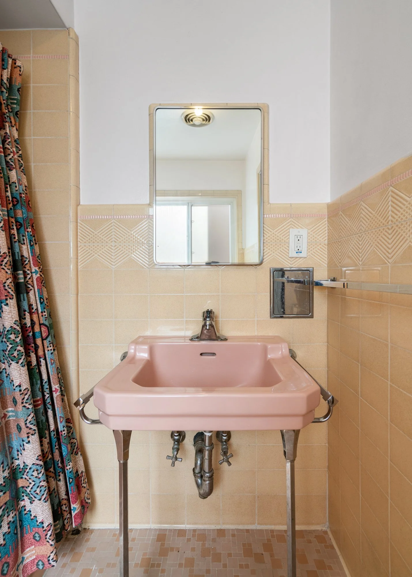 Retro pink baby changing station with metal legs in a vintage bathroom, beige tiled walls, a mirror, a power outlet, and a colorful patterned shower curtain to the left.