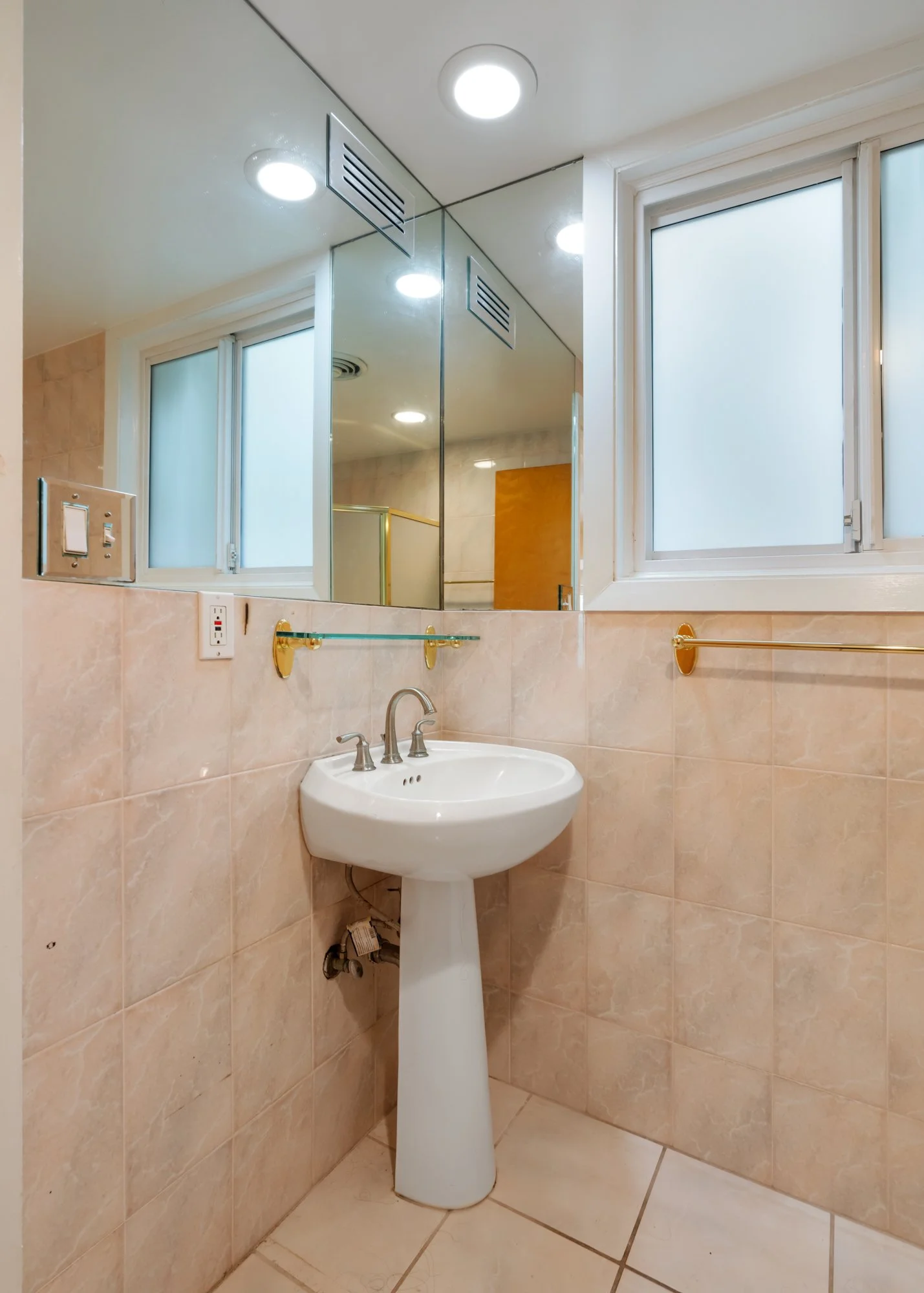 Bathroom with beige tiled walls and floor, a pedestal sink with a silver faucet, gold towel bars, a mirrored medicine cabinet, and a frosted window.