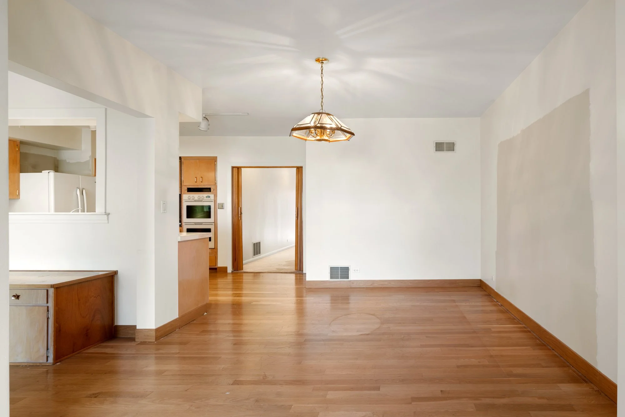 Empty living room with hardwood floors, white walls, a ceiling light fixture, and views into the kitchen and hallway.