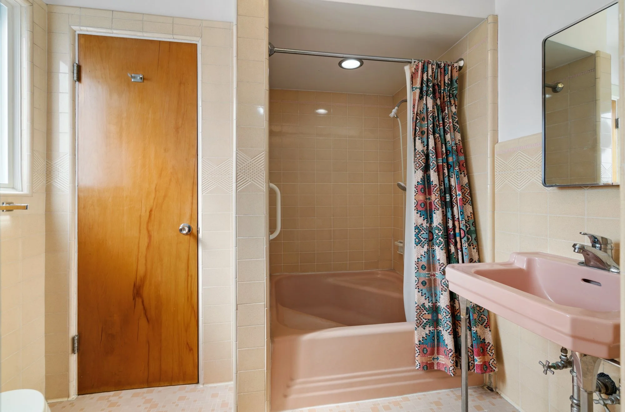 Vintage bathroom with beige tiled walls and floor, pink bathtub with shower curtain, pink sink, wooden door, and a mirror.