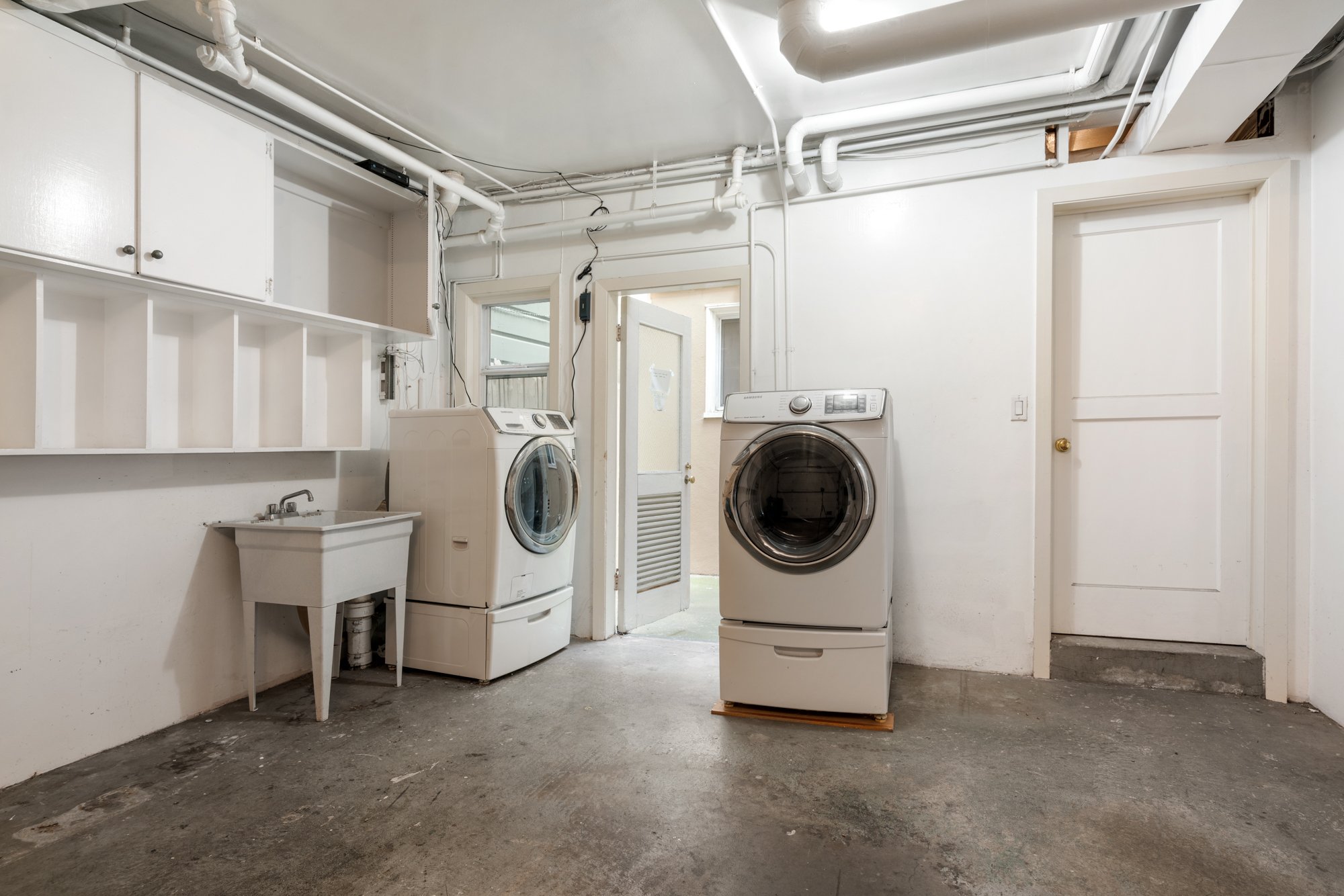 A laundry room with a white washing machine and dryer, a utility sink, white cabinets, and exposed pipes on the ceiling.