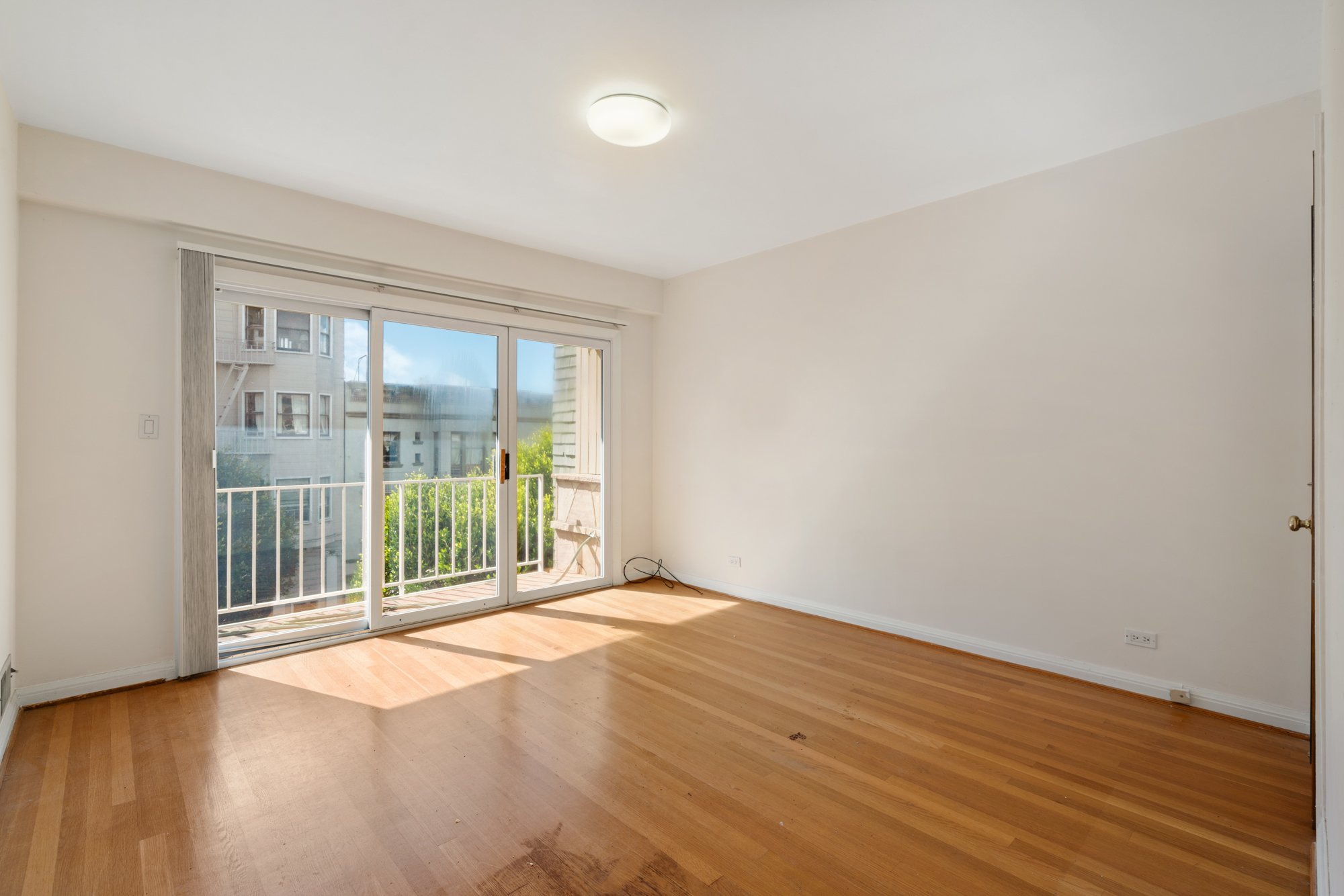 Empty room with hardwood flooring, a sliding glass door leading to a balcony, and a ceiling light fixture.
