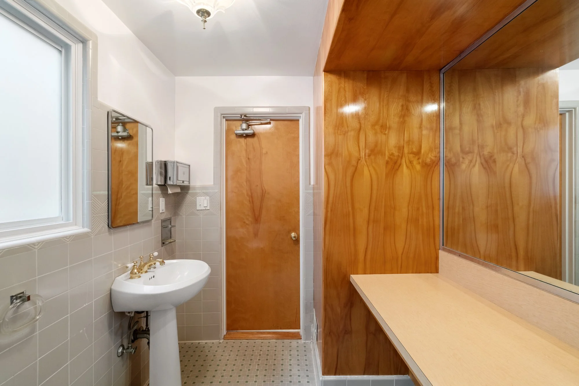 Small bathroom with white tiled walls, a window, a wall-mounted mirror, a pedestal sink with gold faucet, and a wooden cabinet with a large mirror.
