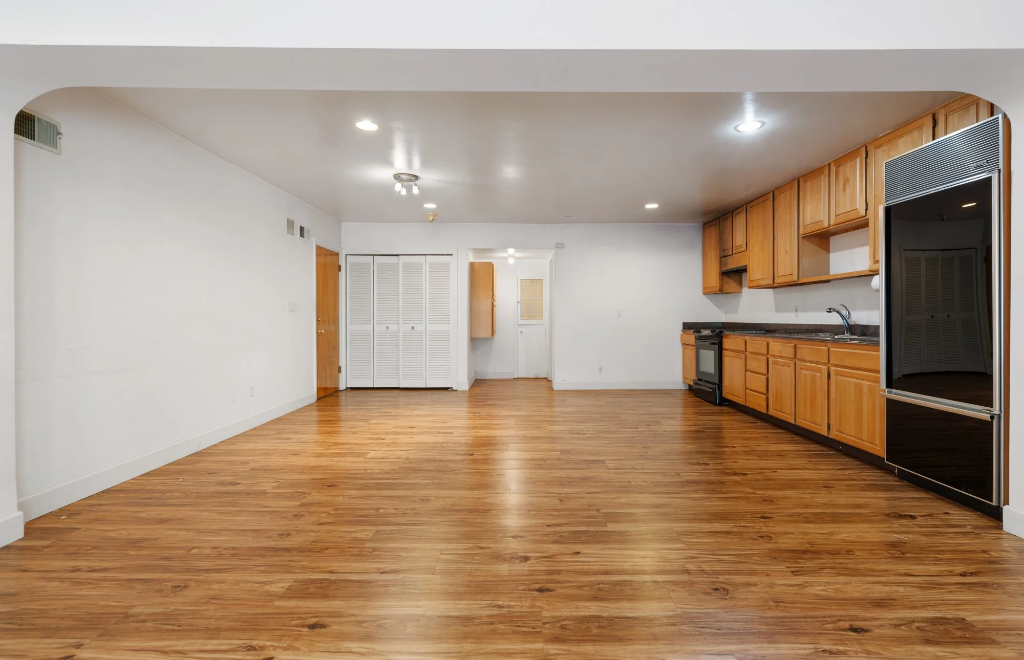 Interior of a room with hardwood floors, white walls, and wooden kitchen cabinets on the right side.