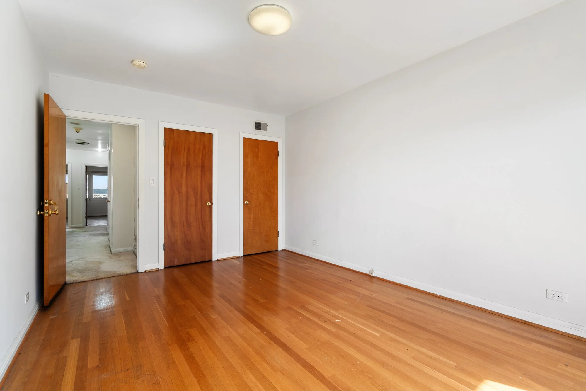 Empty room with hardwood floors, white walls, and wooden doors, with an open doorway showing another room with a window.