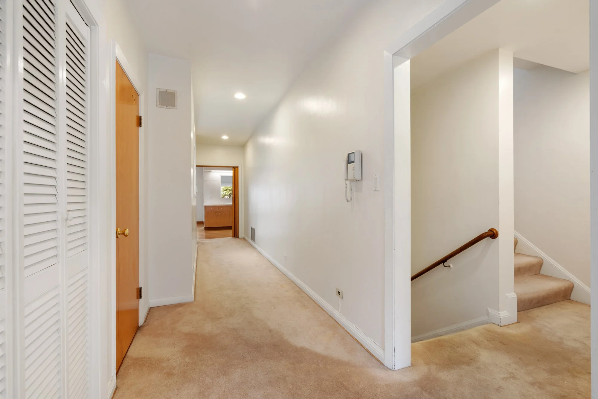 A hallway in a home with beige carpet, white walls, a wooden door on the left, a wall-mounted thermostat, ceiling lights, and a staircase with a wooden handrail leading upward on the right. There is an opening at the end of the hallway revealing a ki