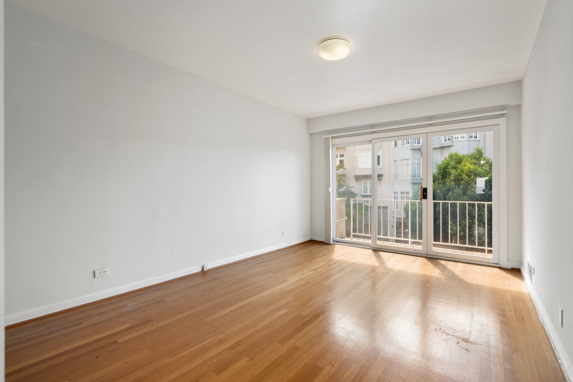 Empty living room with hardwood floors, white walls, and a large glass sliding door leading to a balcony with view of trees and neighboring buildings.