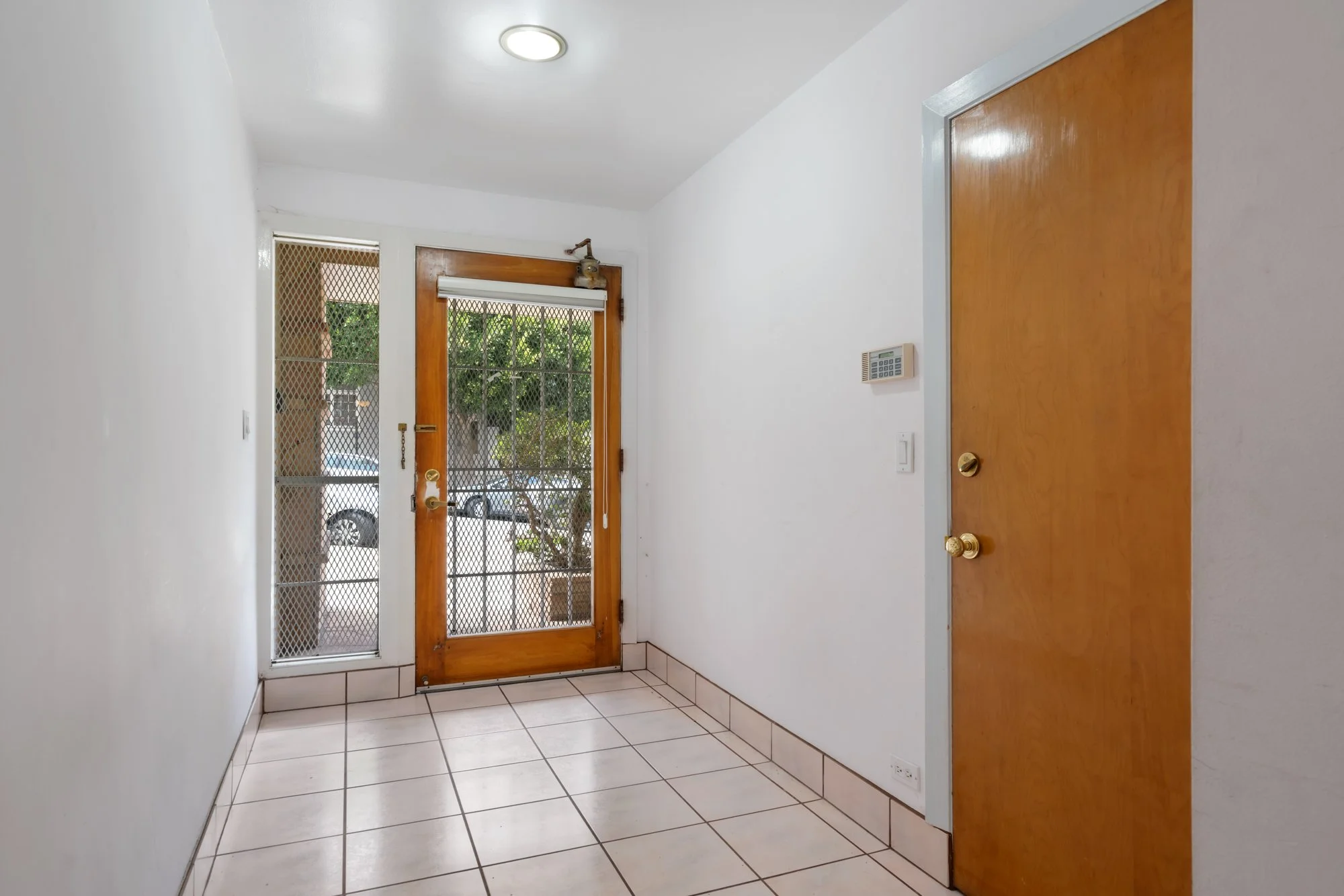 Entryway with white walls, beige tiled floor, wooden door leading outside, security screen door, and a keypad on the wall.