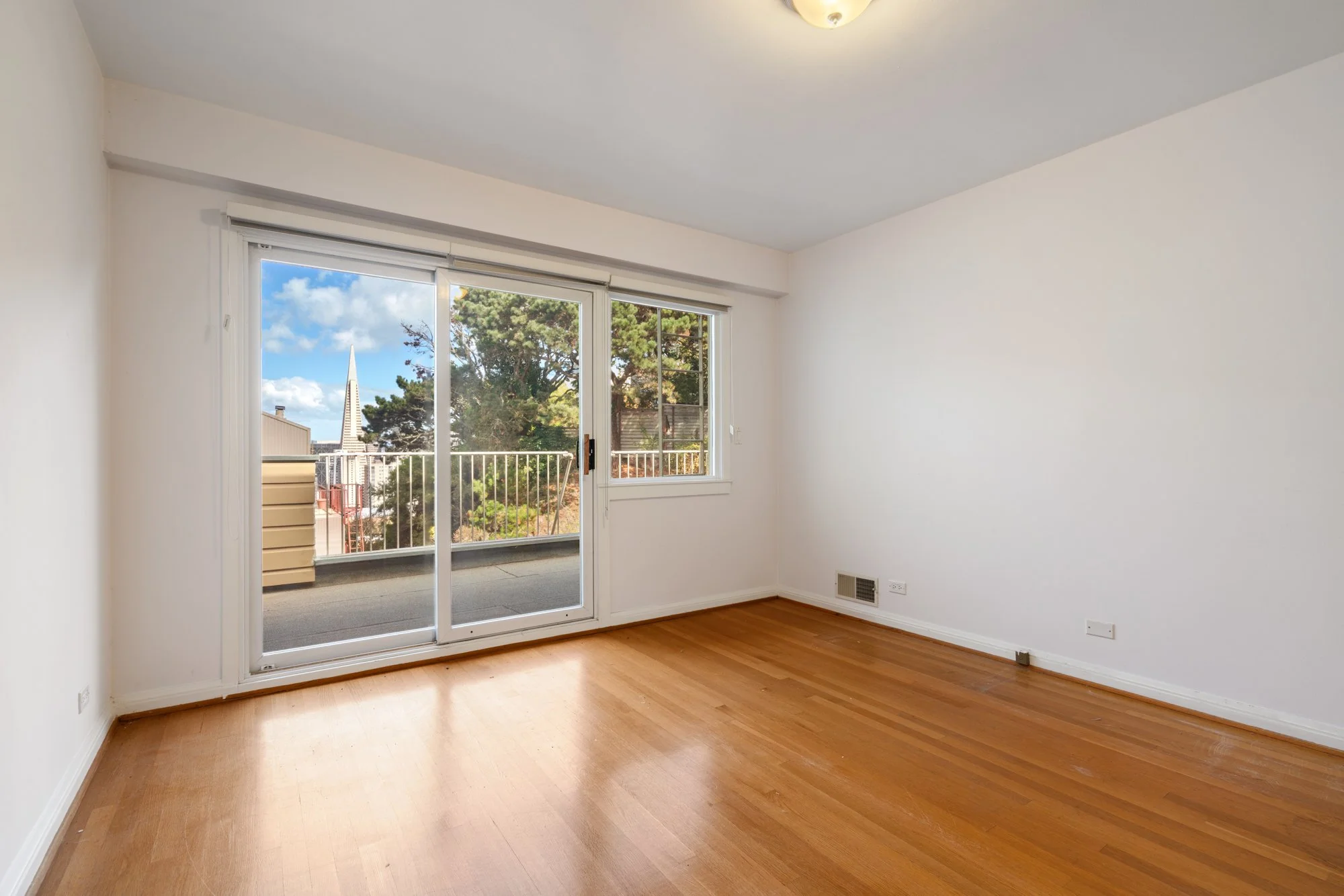 Empty room with hardwood floor and sliding glass door leading to balcony, offering a view of trees and a church steeple outside.