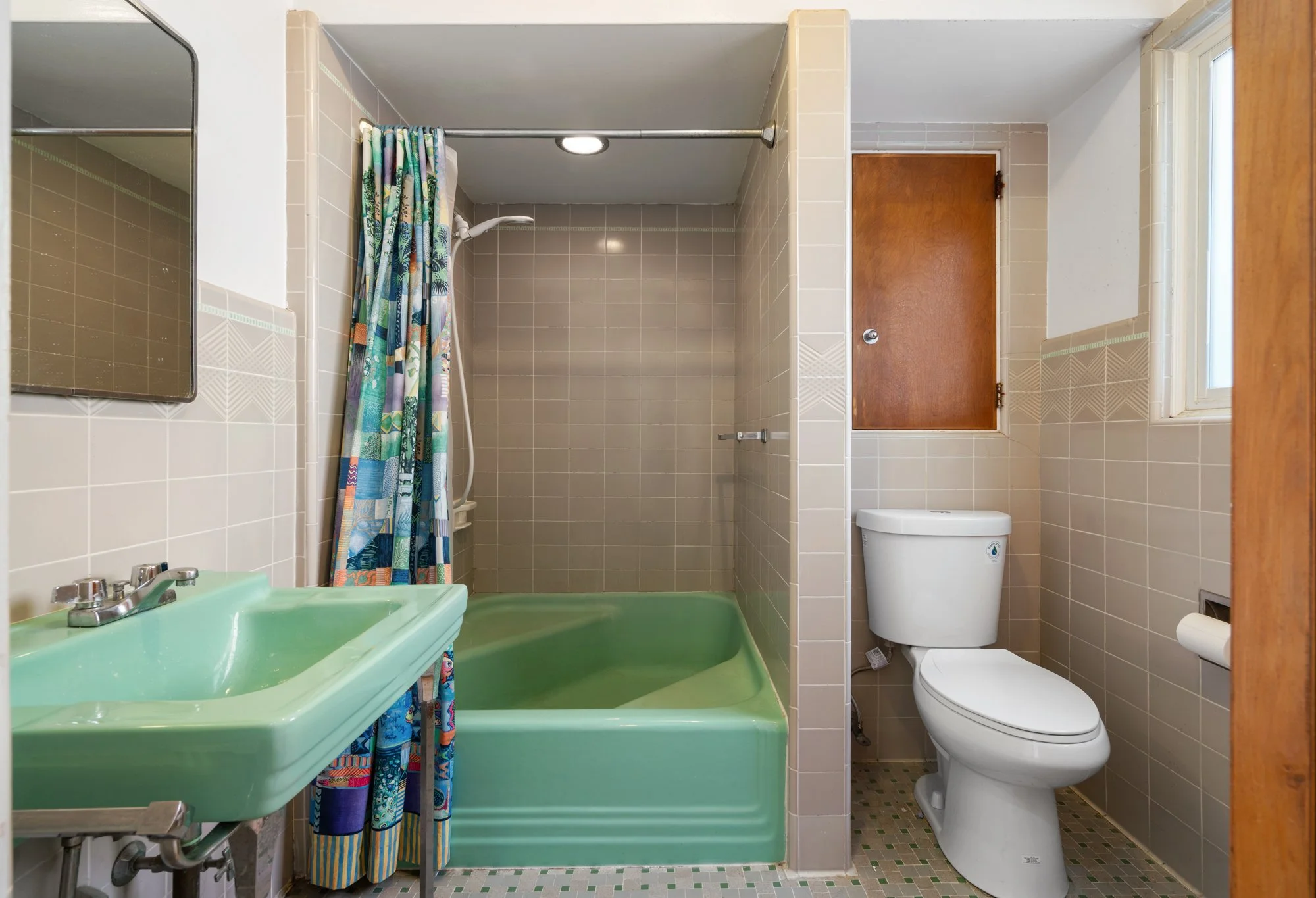 A small bathroom with beige tiled walls and a frosted window. It features a green sink and bathtub, a colorful shower curtain, a wooden cabinet above the toilet, a mirror on the wall, and a patterned tile floor.
