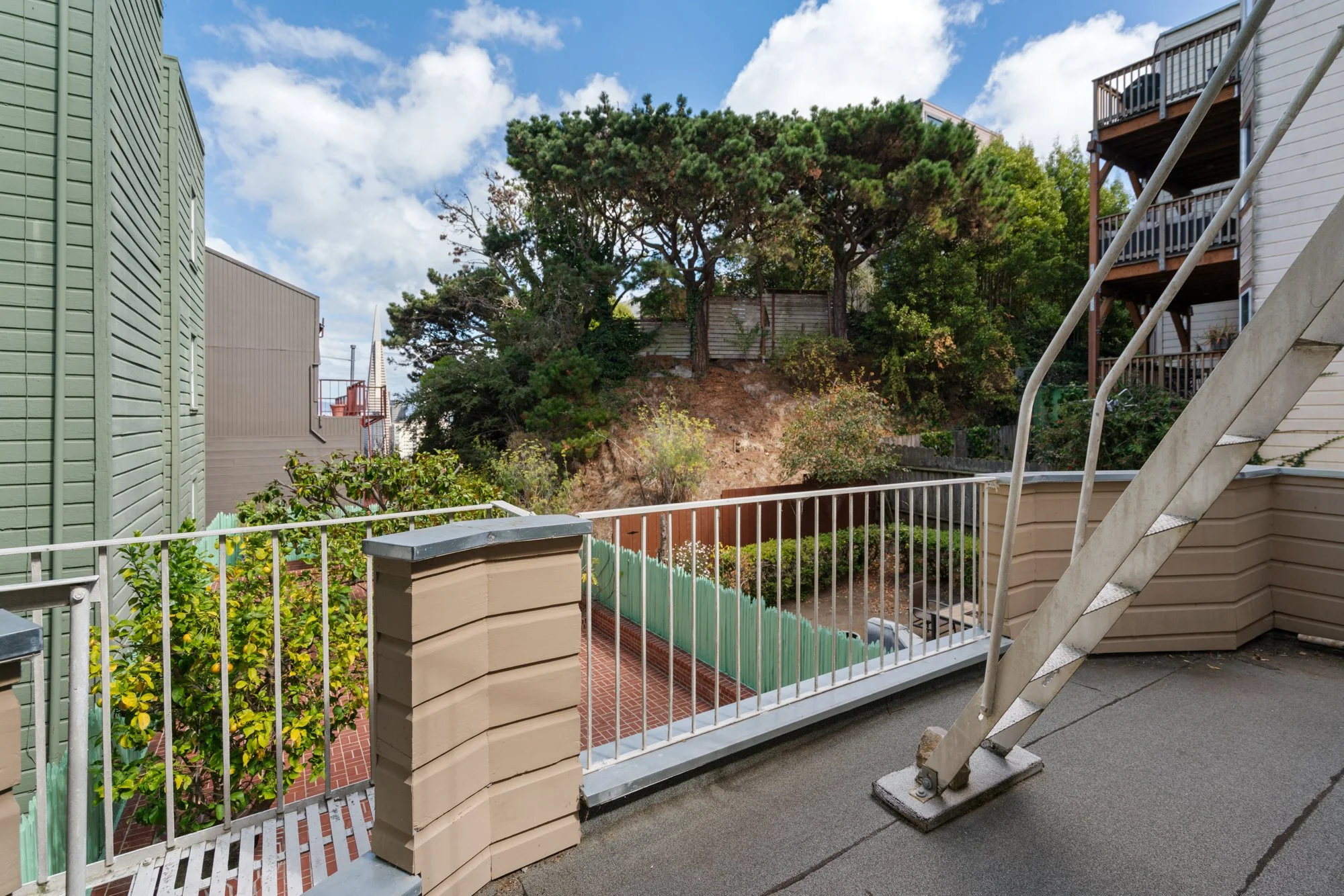View from a balcony showing a staircase, some houses, a backyard with plants, a hillside with trees, a fence, and a partly cloudy sky.